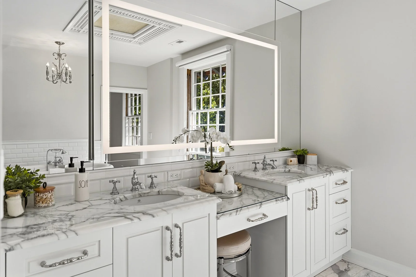 A modern bathroom with white cabinetry, marble countertops, a large illuminated mirror, and a window with a view of green trees outside.