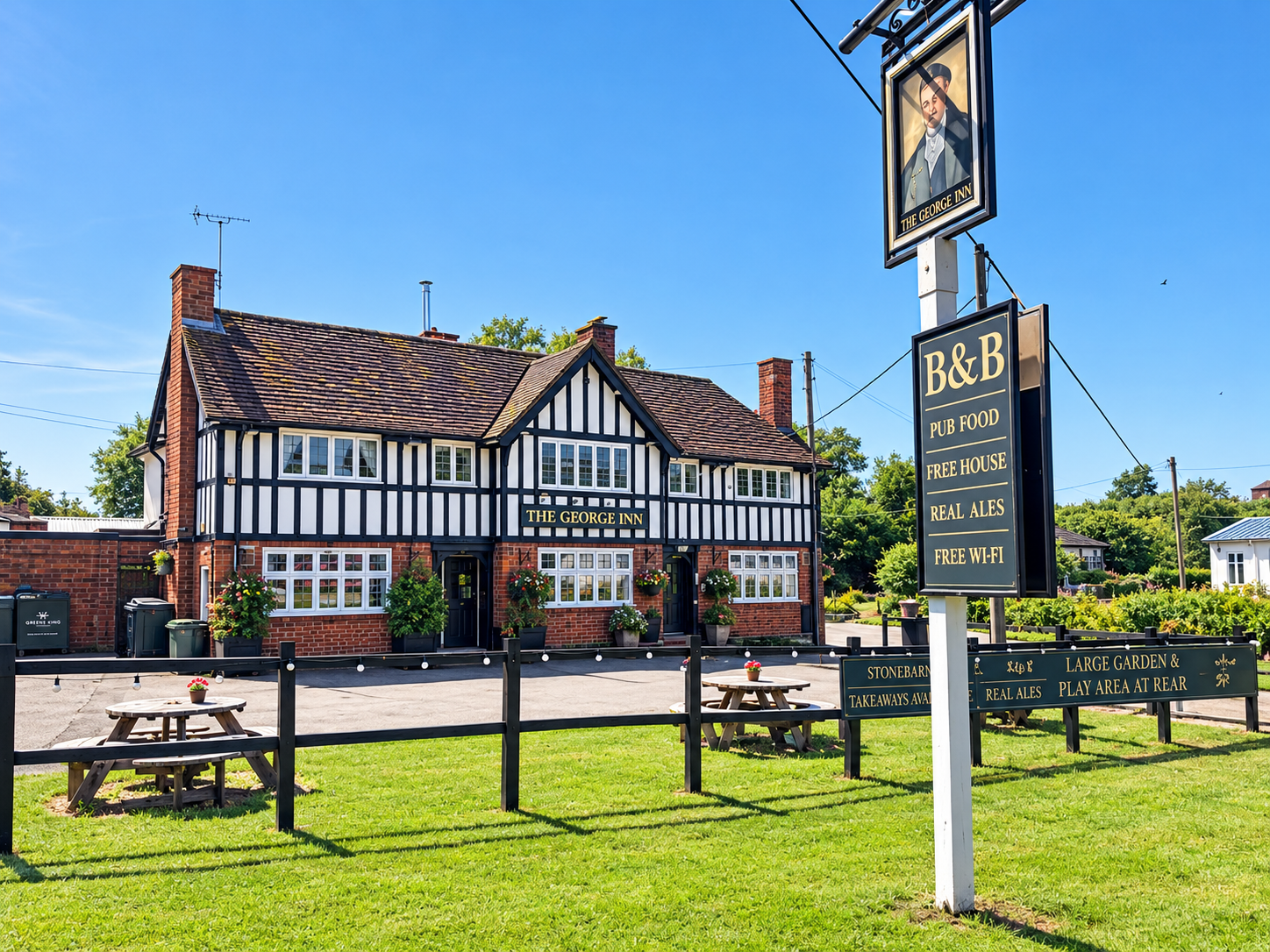 Exterior of The George Inn, a traditional pub with black and white timber framing, brick lower walls, and hanging flower baskets. There are outdoor tables on a grassy area, and a sign listing amenities such as pub food, free house, real ales, and free Wi-Fi, set against a bright blue sky.