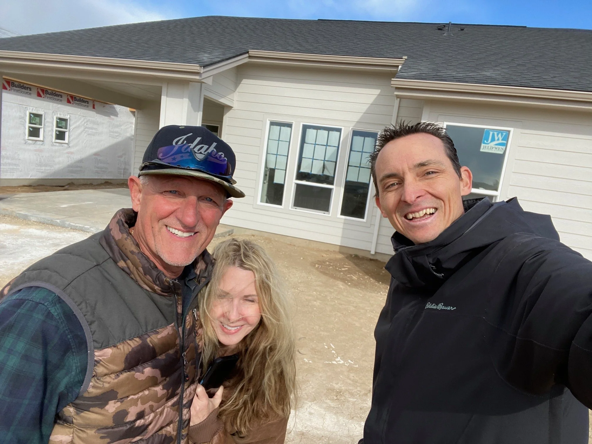 Two smiling men and a woman posing in front of a new house under construction, with some construction signs visible.