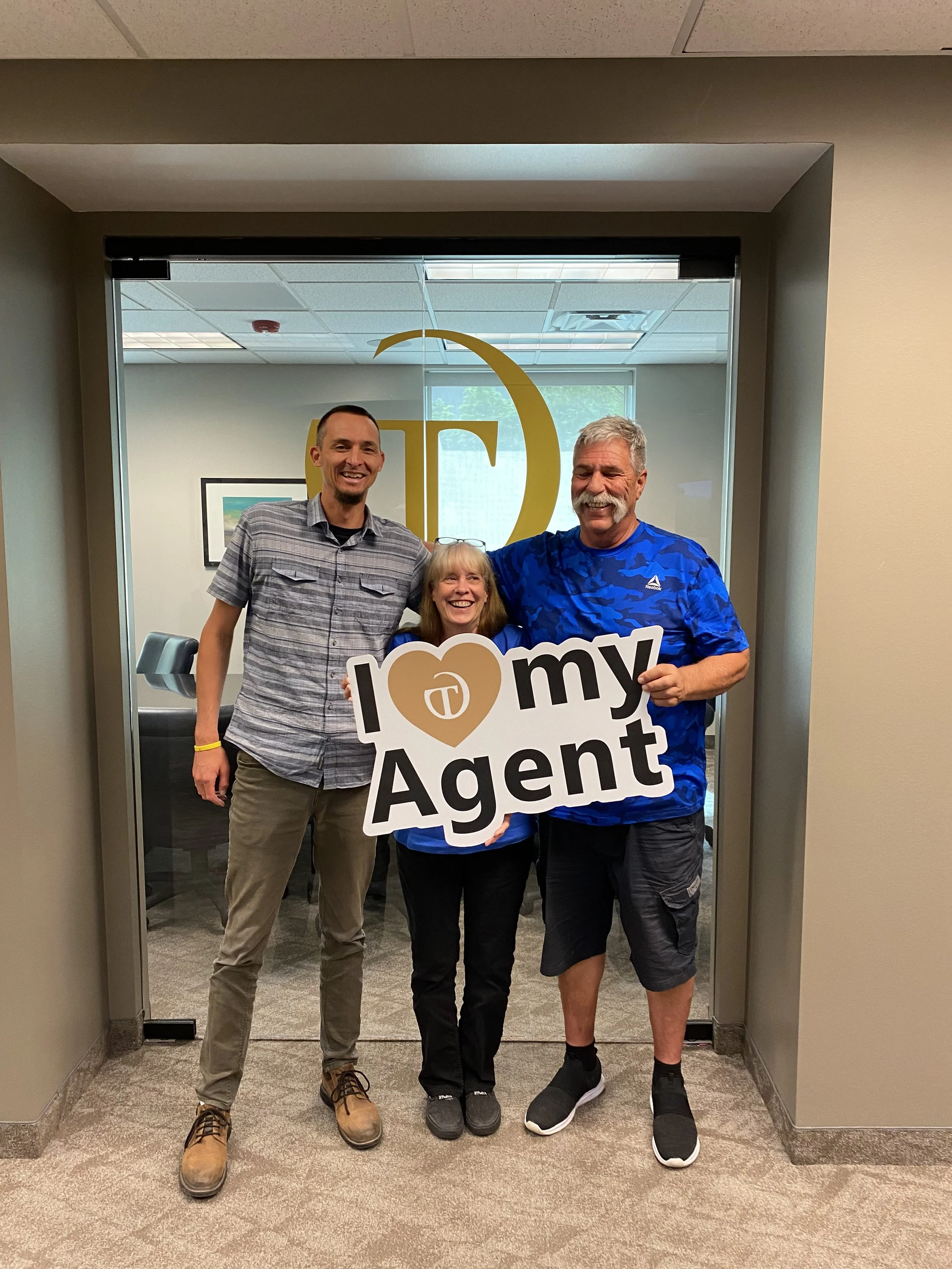 Three smiling people standing together in an office doorway, holding a sign that reads 'I love my agent,' with a glass door behind them and office decor visible inside.