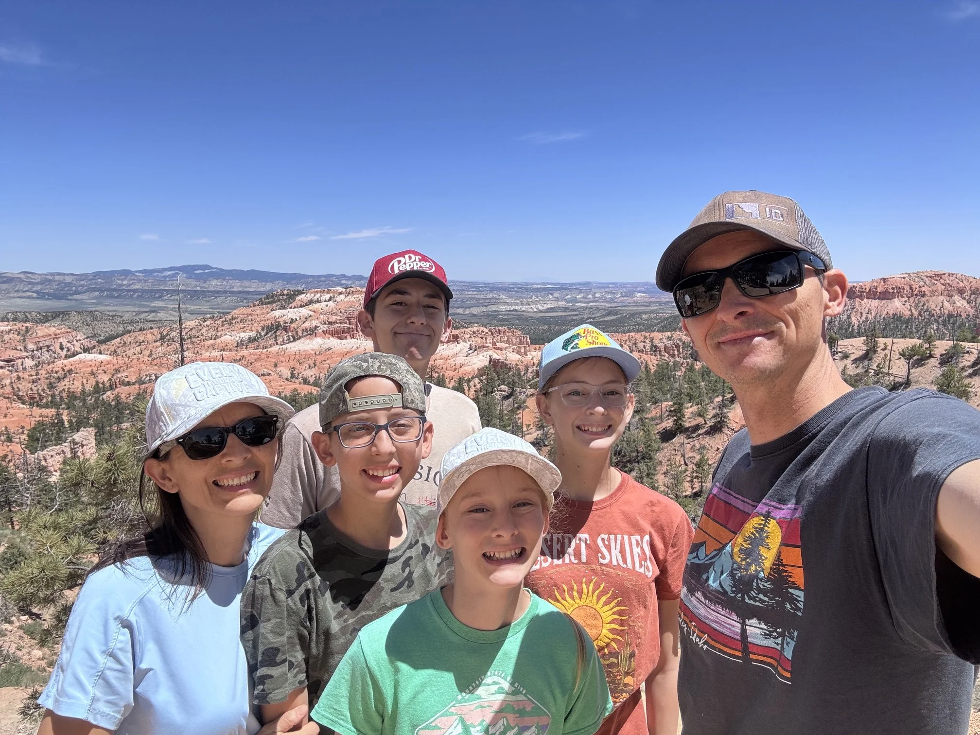A group of seven people, including children and an adult, posing for a selfie with a scenic desert landscape featuring orange rock formations and a clear blue sky in the background.