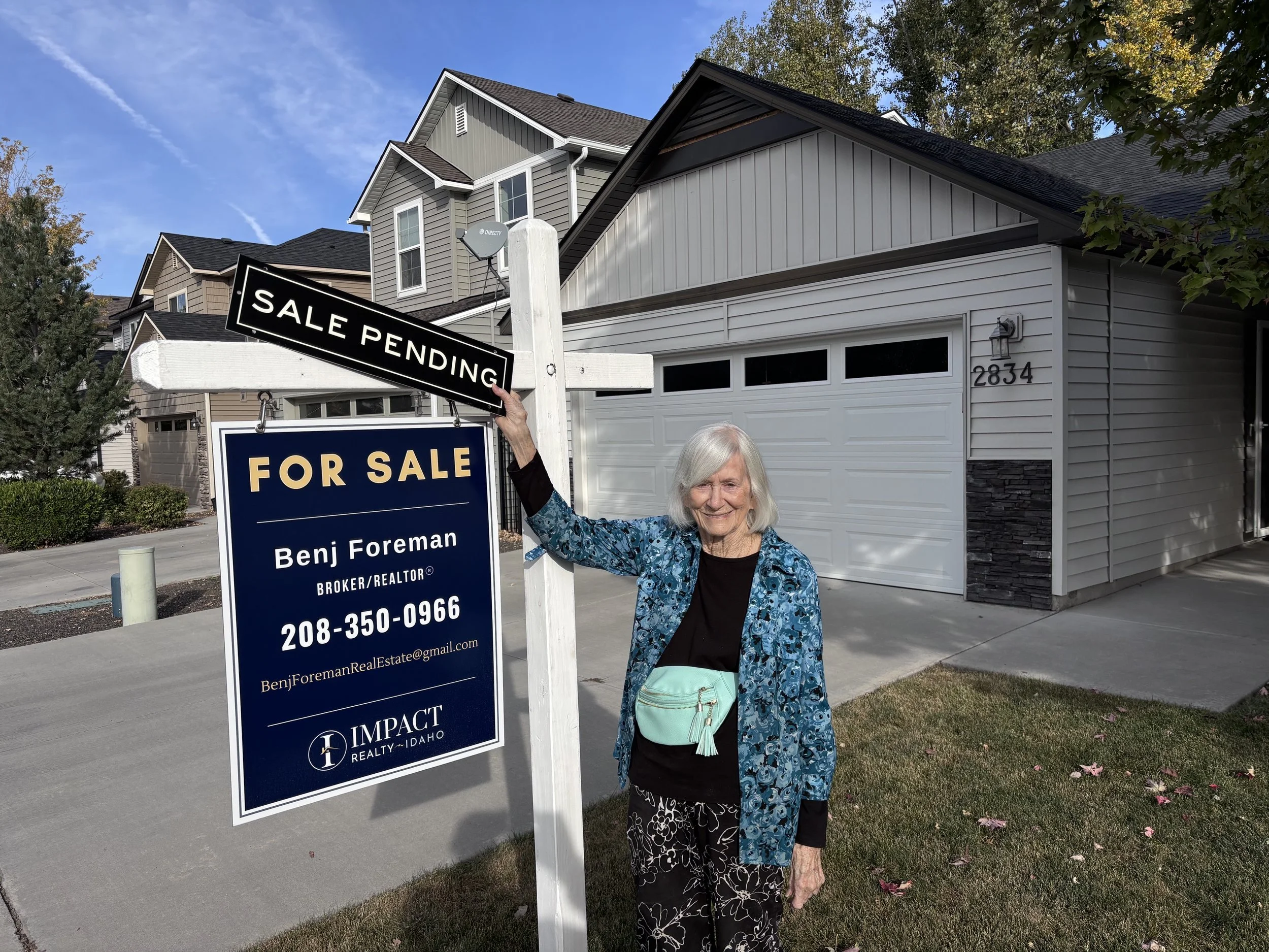An elderly woman standing in front of a house with a "Sale Pending" sign and a "For Sale" sign, holding the "Sale Pending" sign. The house has gray and white exterior with a garage and the address 2834 visible. The sky is clear and blue with some clo