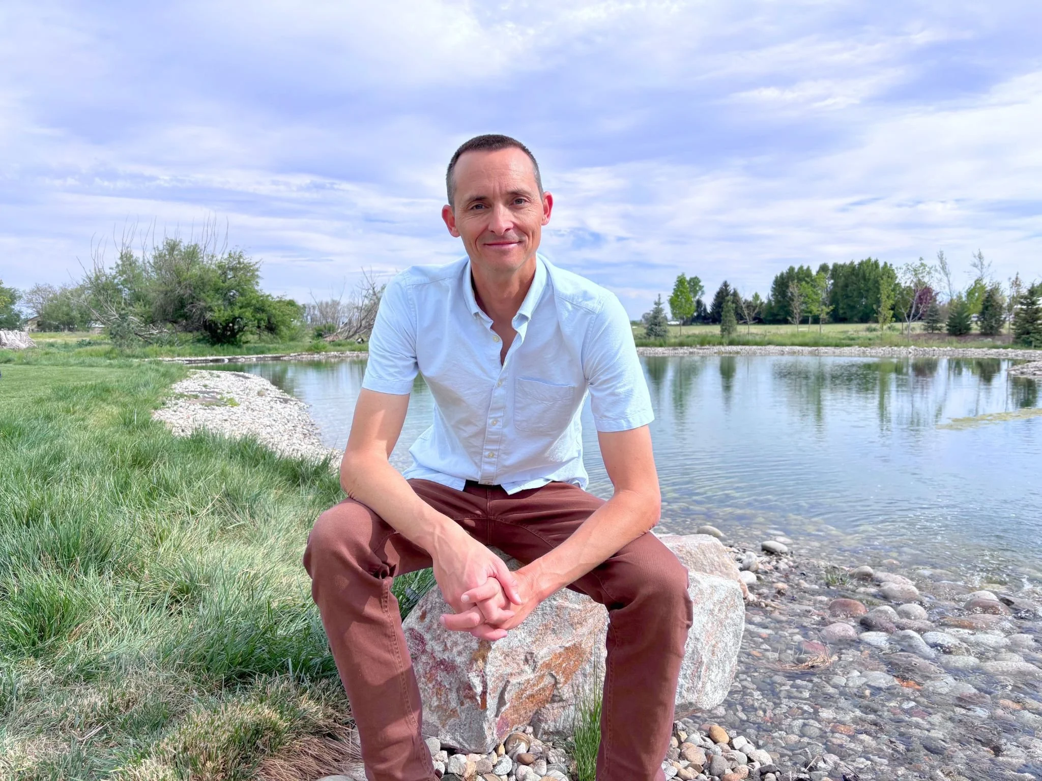 A man in a white shirt and brown pants sitting on a large rock beside a calm river, with green trees and bushes in the background under a partly cloudy sky.