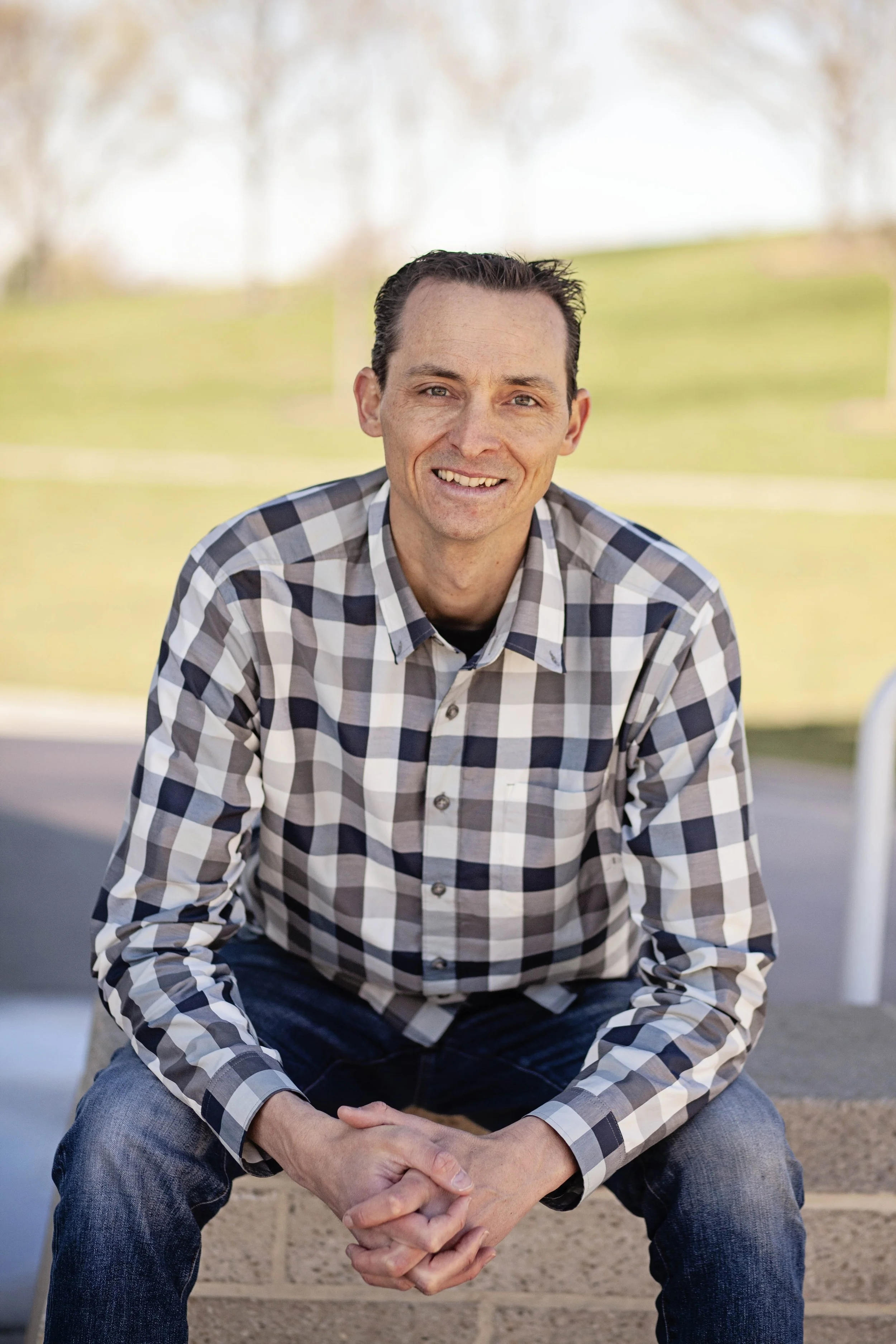 A man in a plaid shirt sitting outside, smiling with park trees in the background.