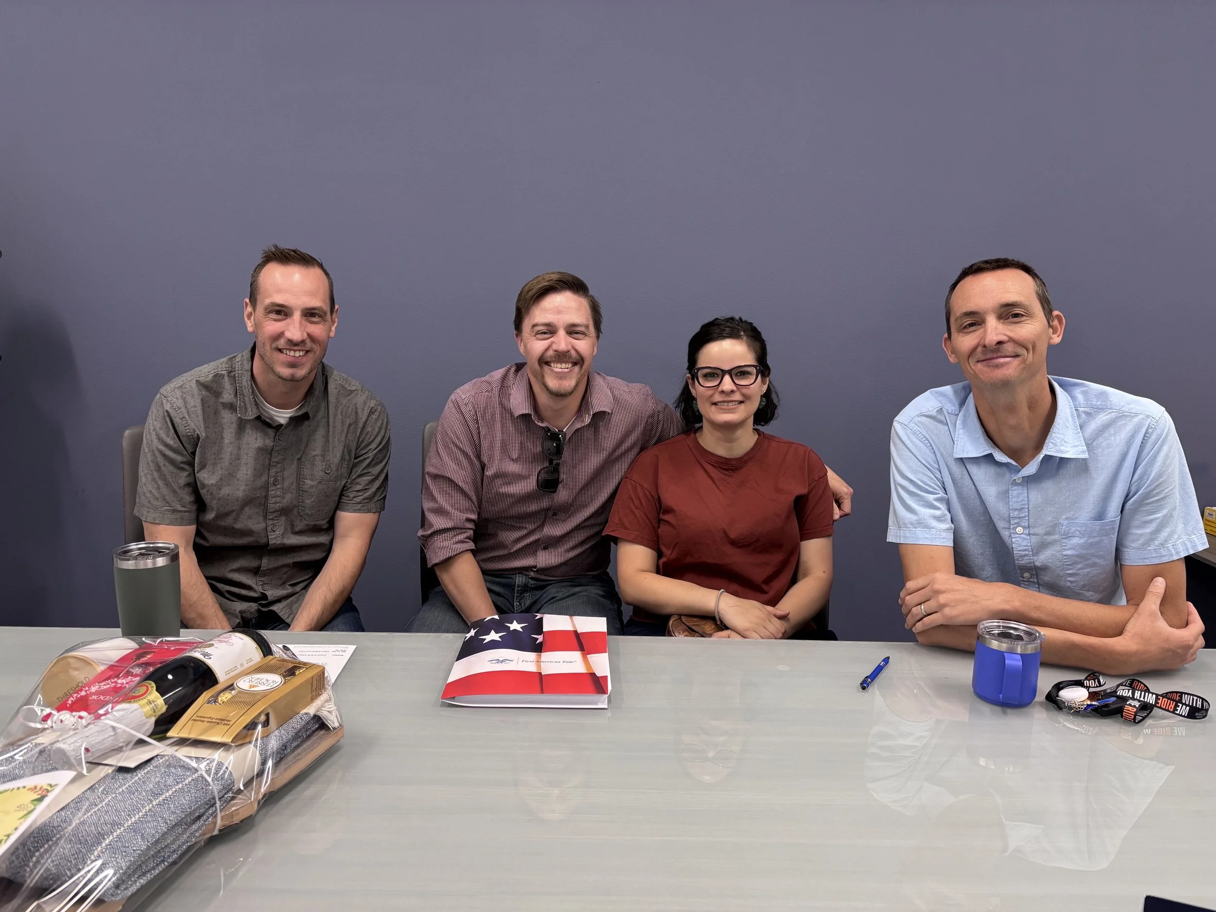 Four people sitting at a table, smiling, with snacks, drinks, and American flags in front of them, against a plain blue wall background.