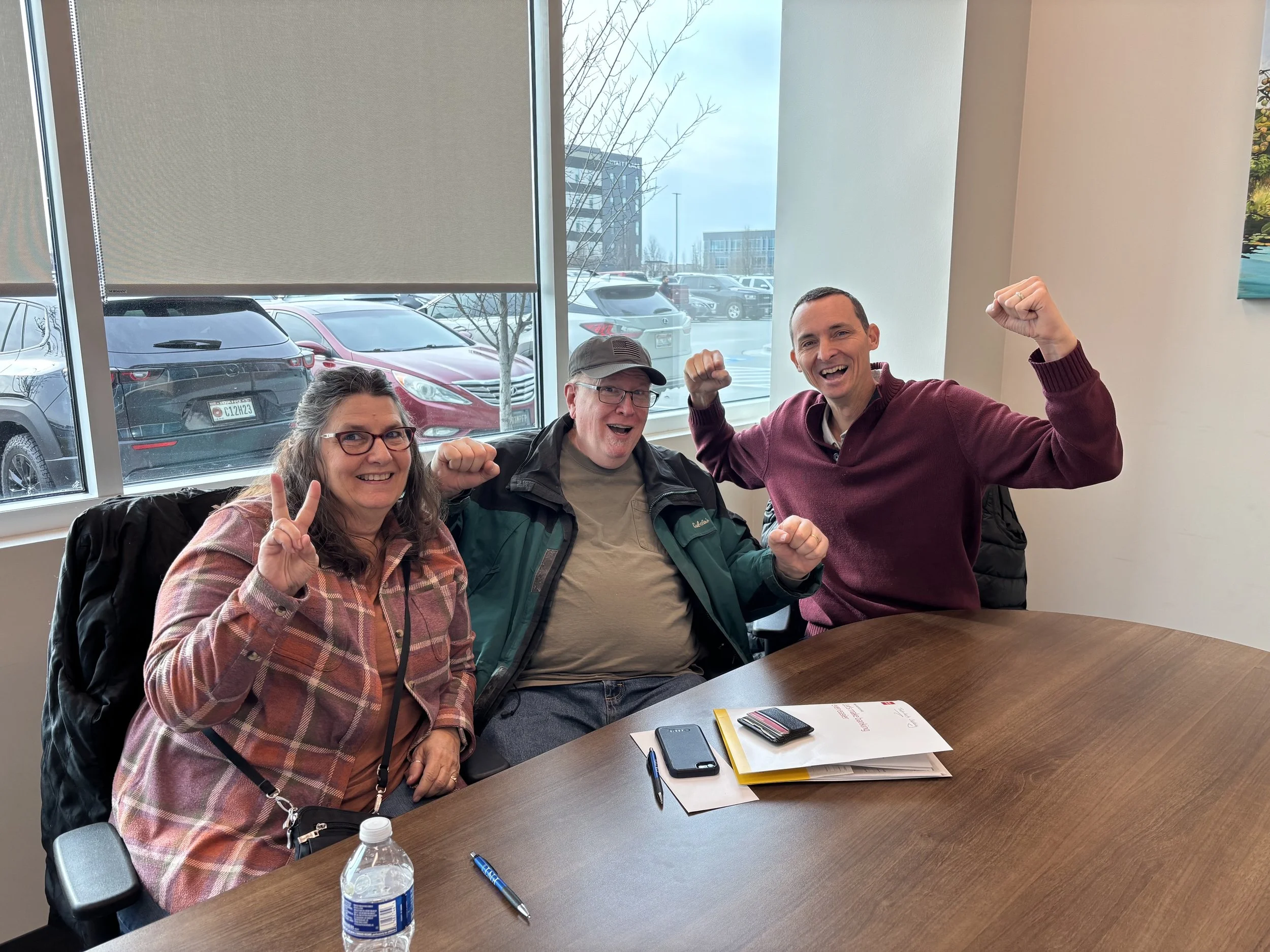 Three people sitting at a wooden table in a meeting room, smiling and raising their fists in celebration. The table has a water bottle, papers, a pen, and a phone. Large window behind them shows parked cars and buildings outside.