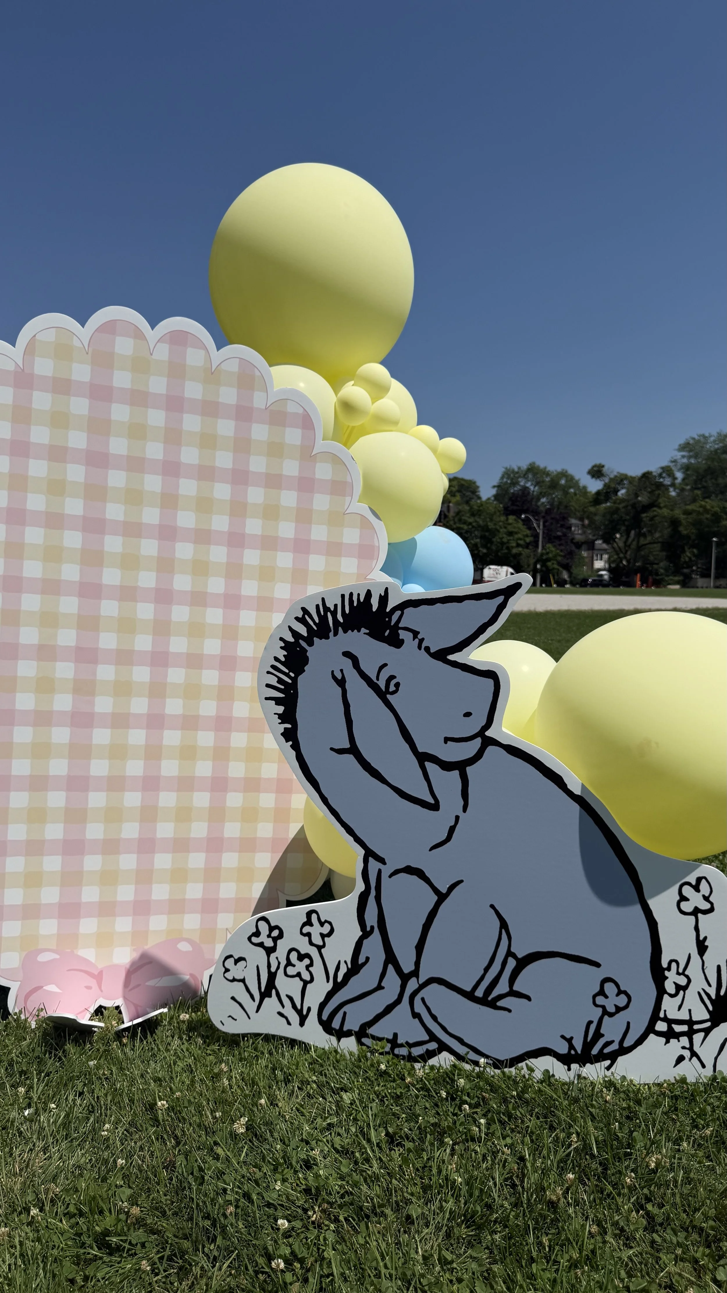 Colorful balloons, a pink checkered backdrop, and a black-and-white cartoon cutout of a person with antlers, sitting with their hand on their face, amid small flowers, outdoors on a sunny day.