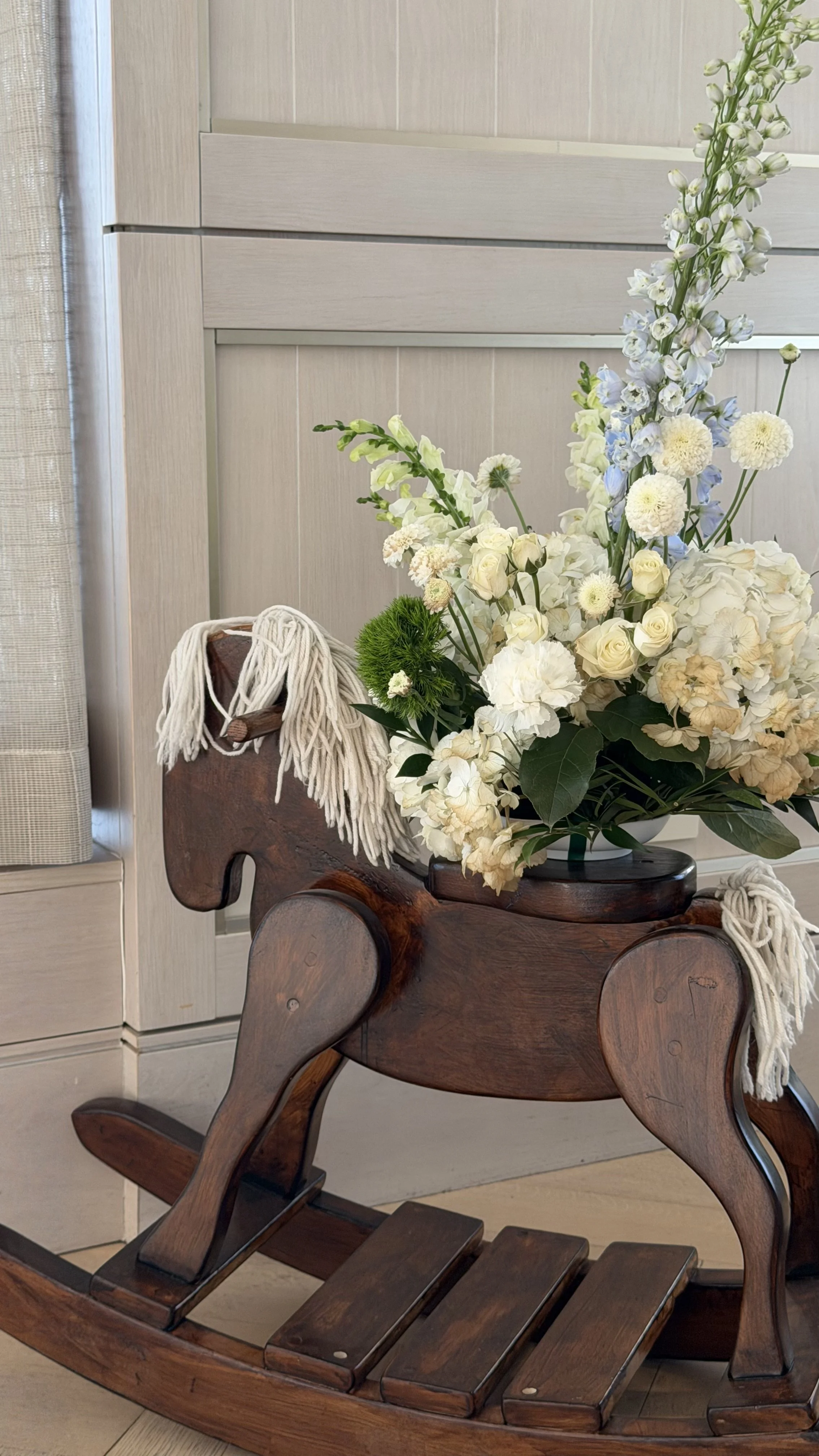 A wooden rocking horse with a bouquet of white and light blue flowers placed on it.