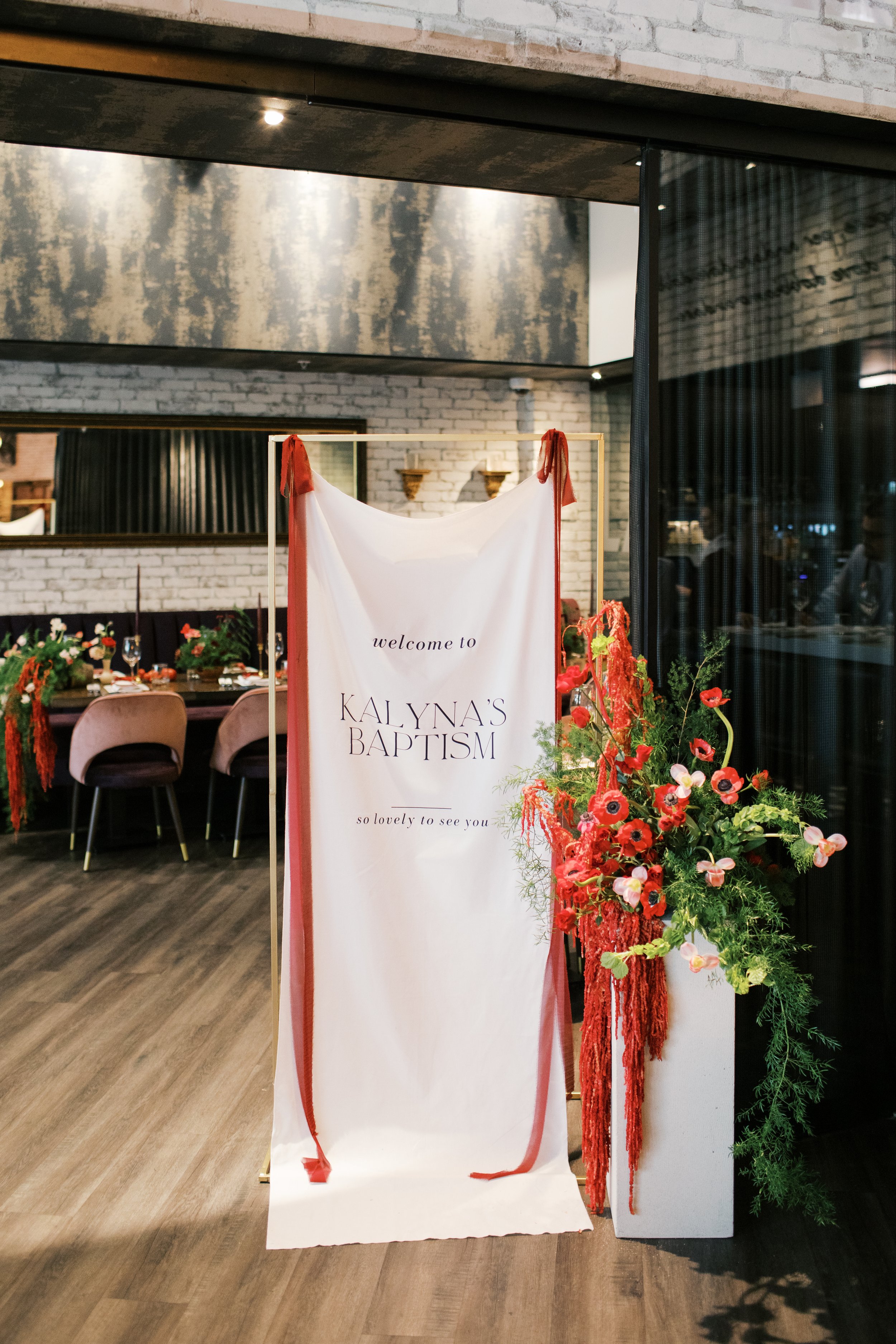 A white fabric backdrop with red trim and the words 'welcome to KALYNA'S BAPTISM so lovely to see you' written on it, accompanied by a floral arrangement of red and pink flowers and greenery, set up in a decorated indoor space with tables, chairs, and rustic brick walls.