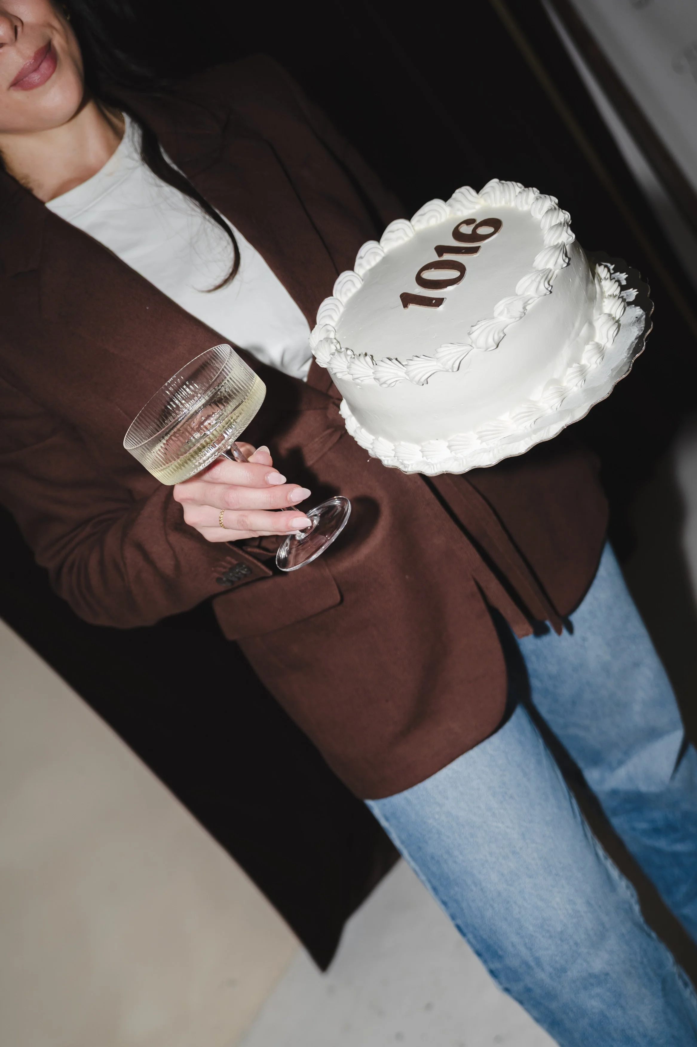 Person holding a white frosted cake with '1016' written in brown icing, decorated with piped white frosting, and a wine glass in their hand, wearing a brown blazer and white shirt.