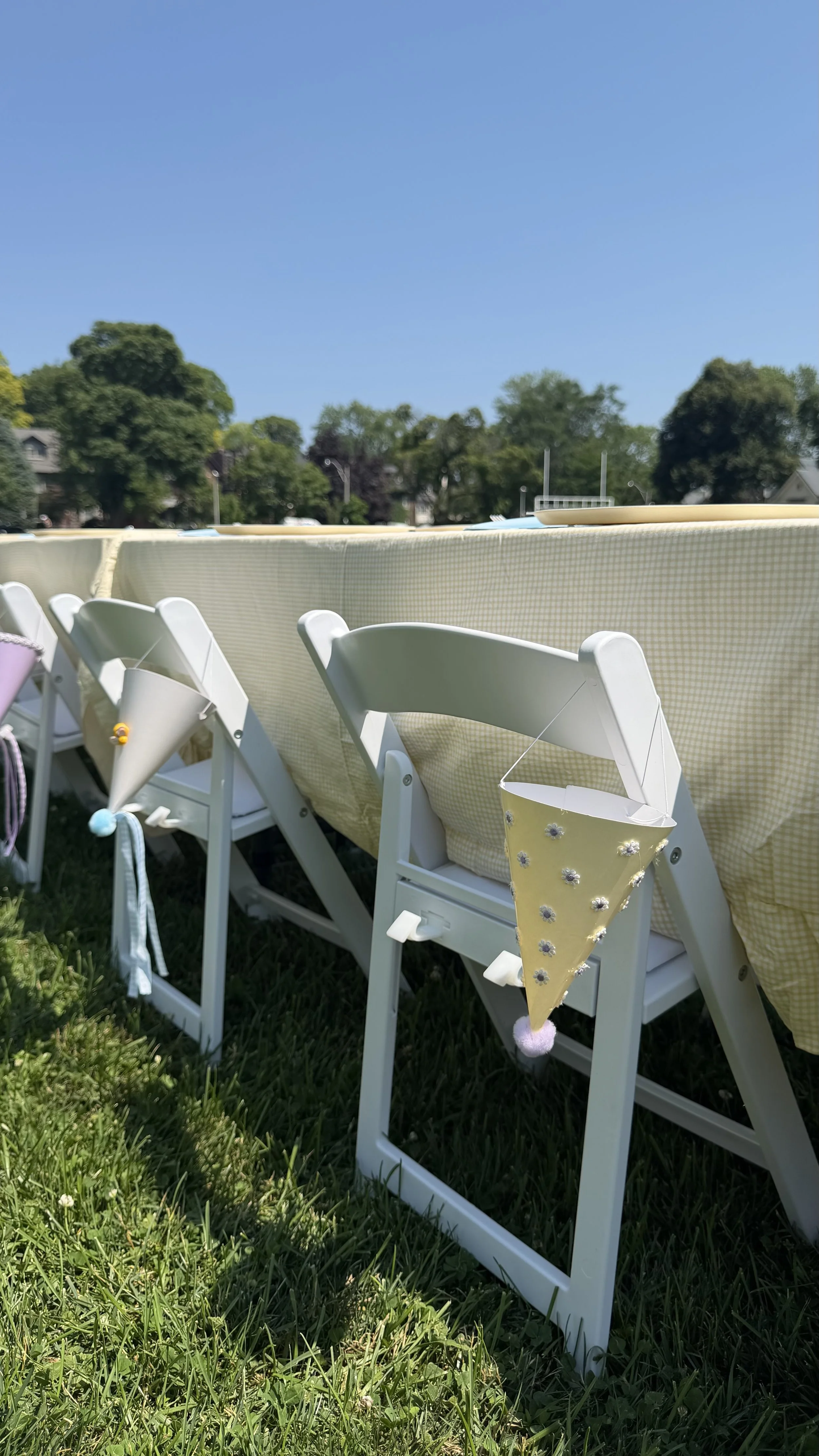 Outdoor scene with green grass, white folding chairs, and decorations for a celebration, against a background of trees and blue sky.