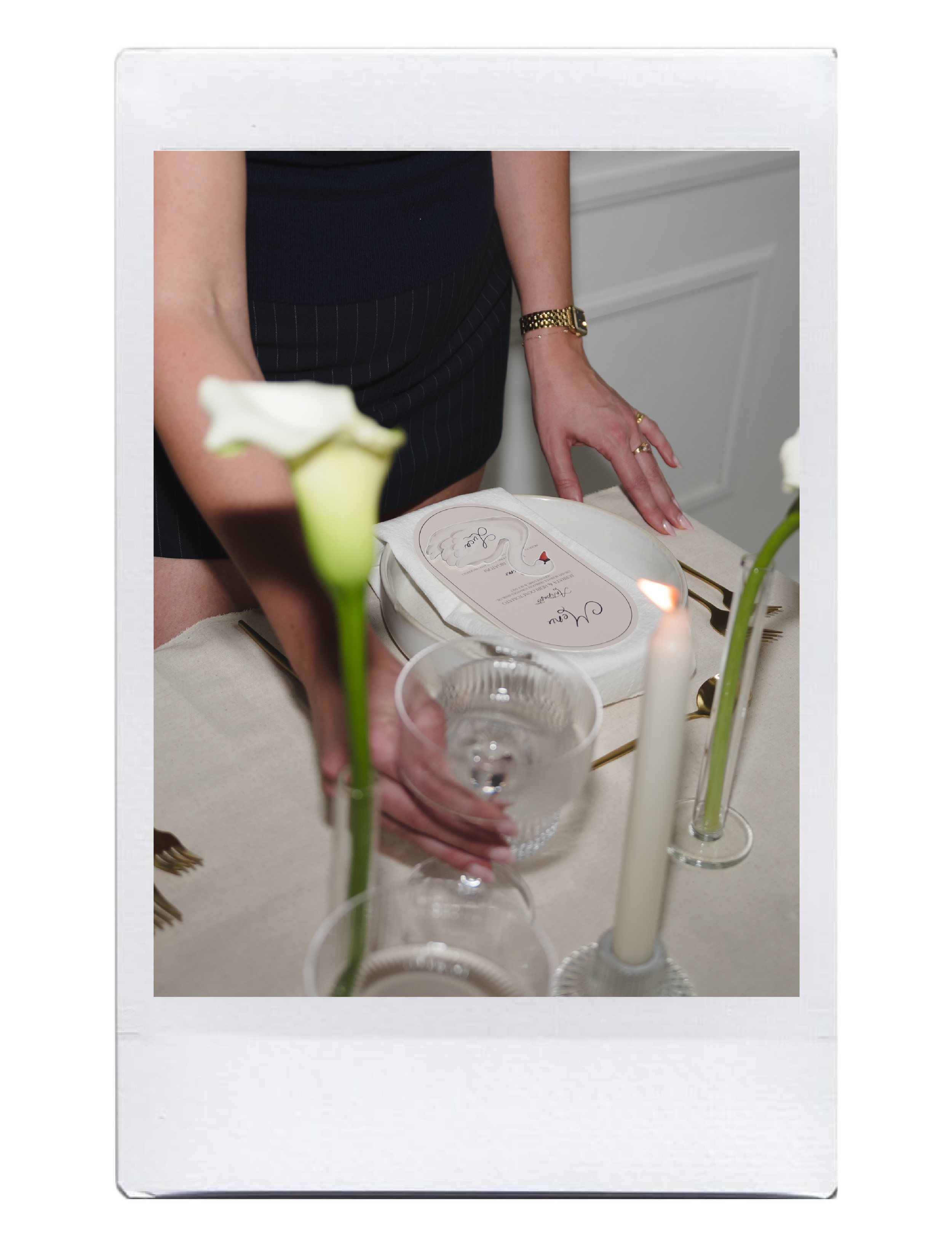 A person wearing a black top and striped skirt, with a gold watch and rings, is leaning over a table with a cake, flowers, candles, and a glass of water, seen through a mirror frame.