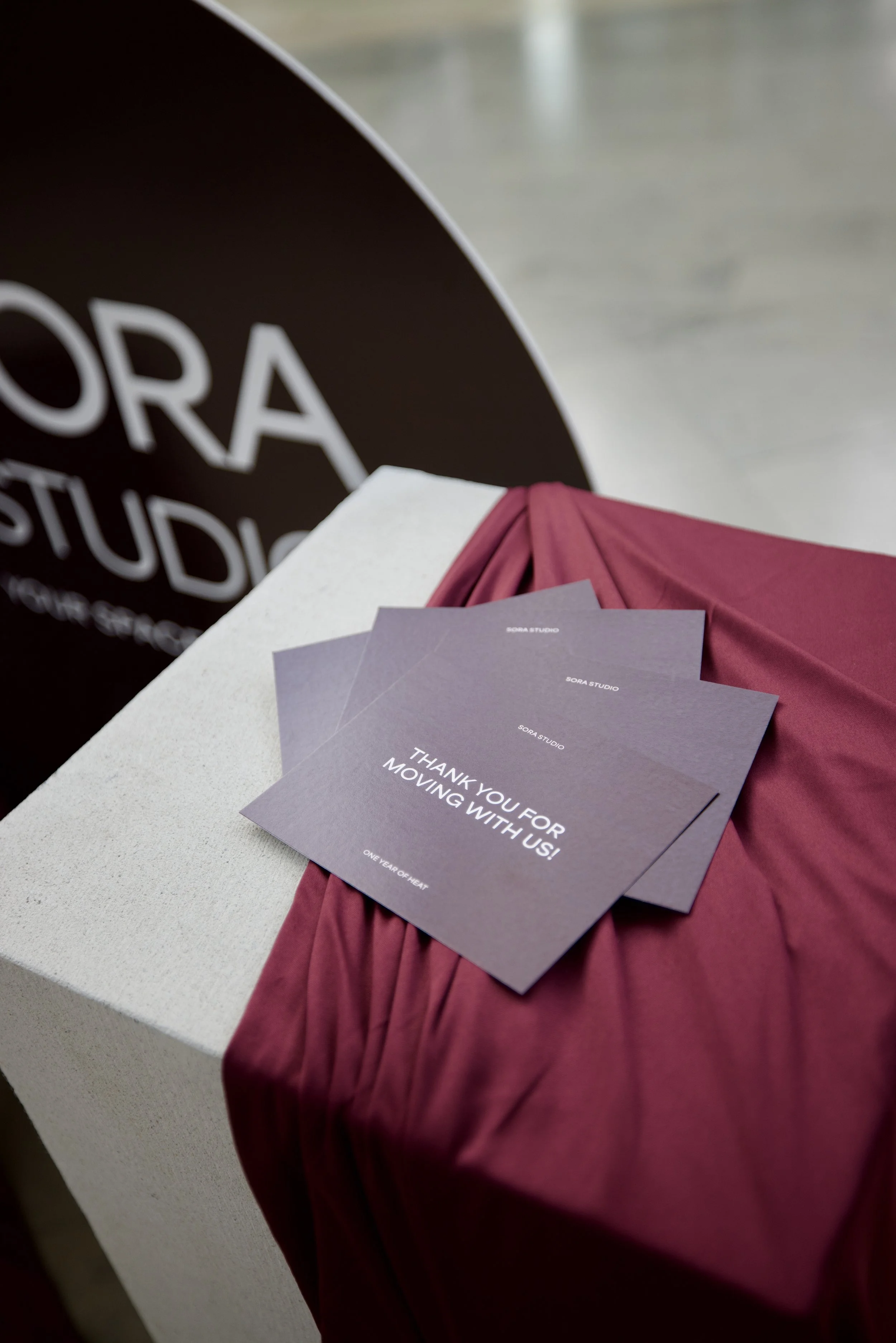 Close-up of purple thank you cards on a table, with pink fabric underneath, and a background with a black and white sign reading 'SORA STUDI'.
