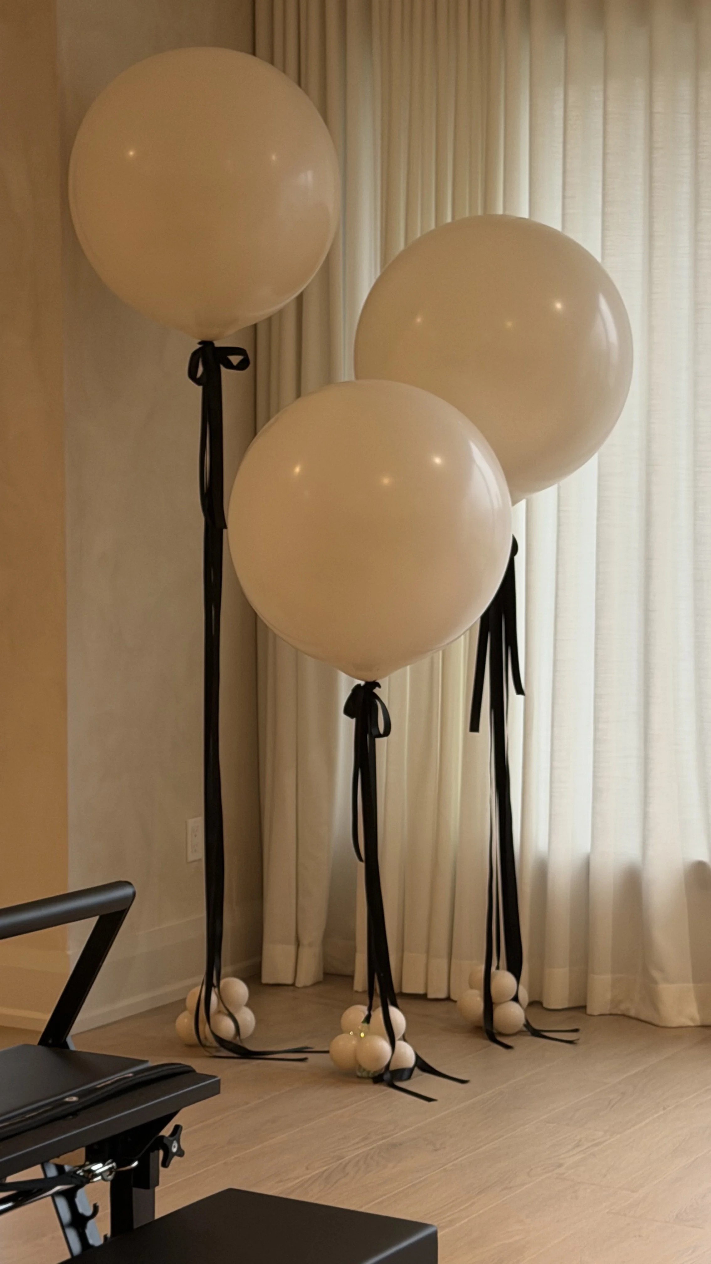 A cluster of three large cream-colored balloons tied with black ribbons, each balloon is attached to multiple smaller white balloons at the bottom, set against beige curtains and a wall in the background.