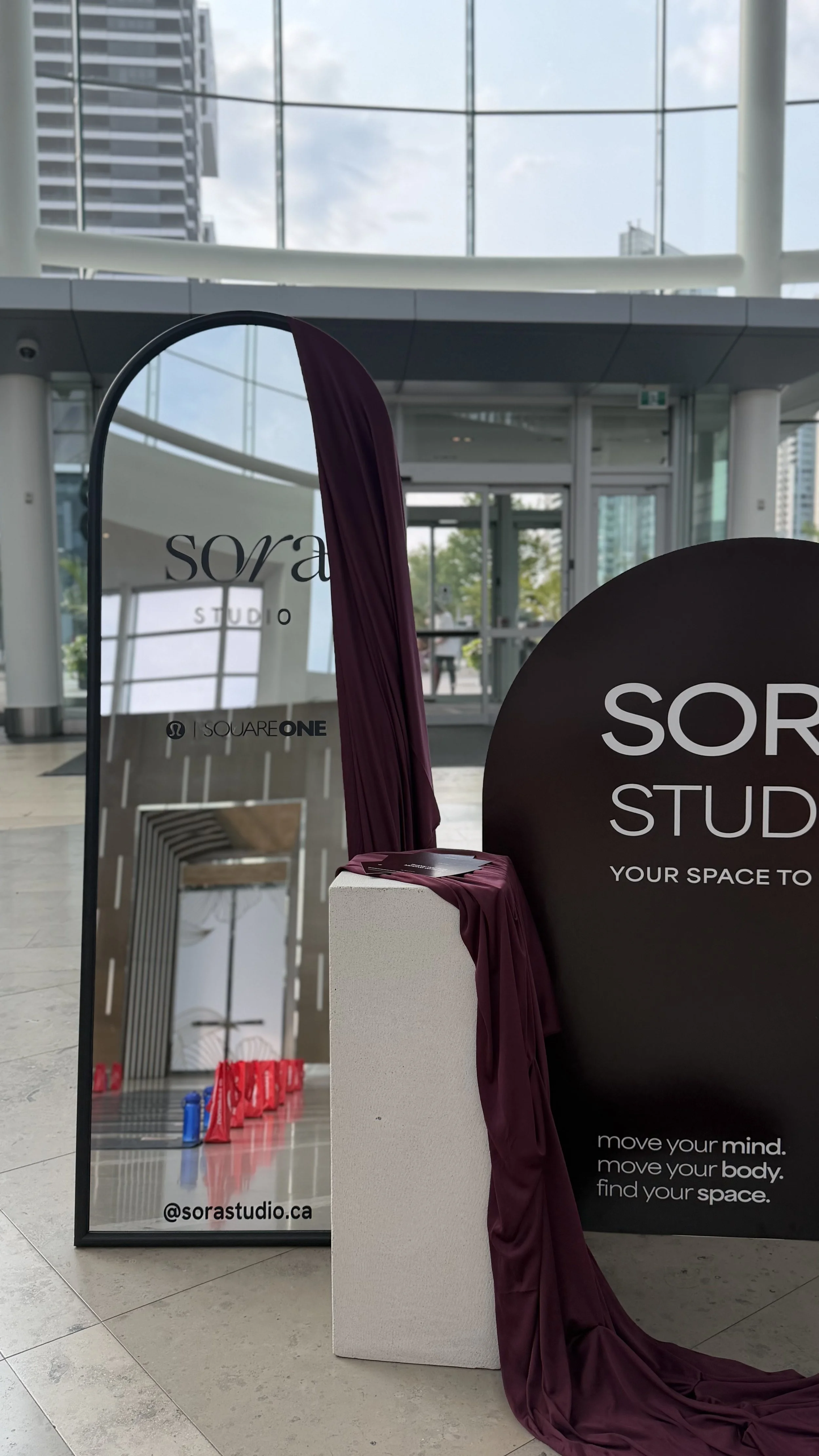 A display booth for Sorra Studio featuring a black sign, reflective mirror, and draped purple fabric inside a modern, glass-walled building.
