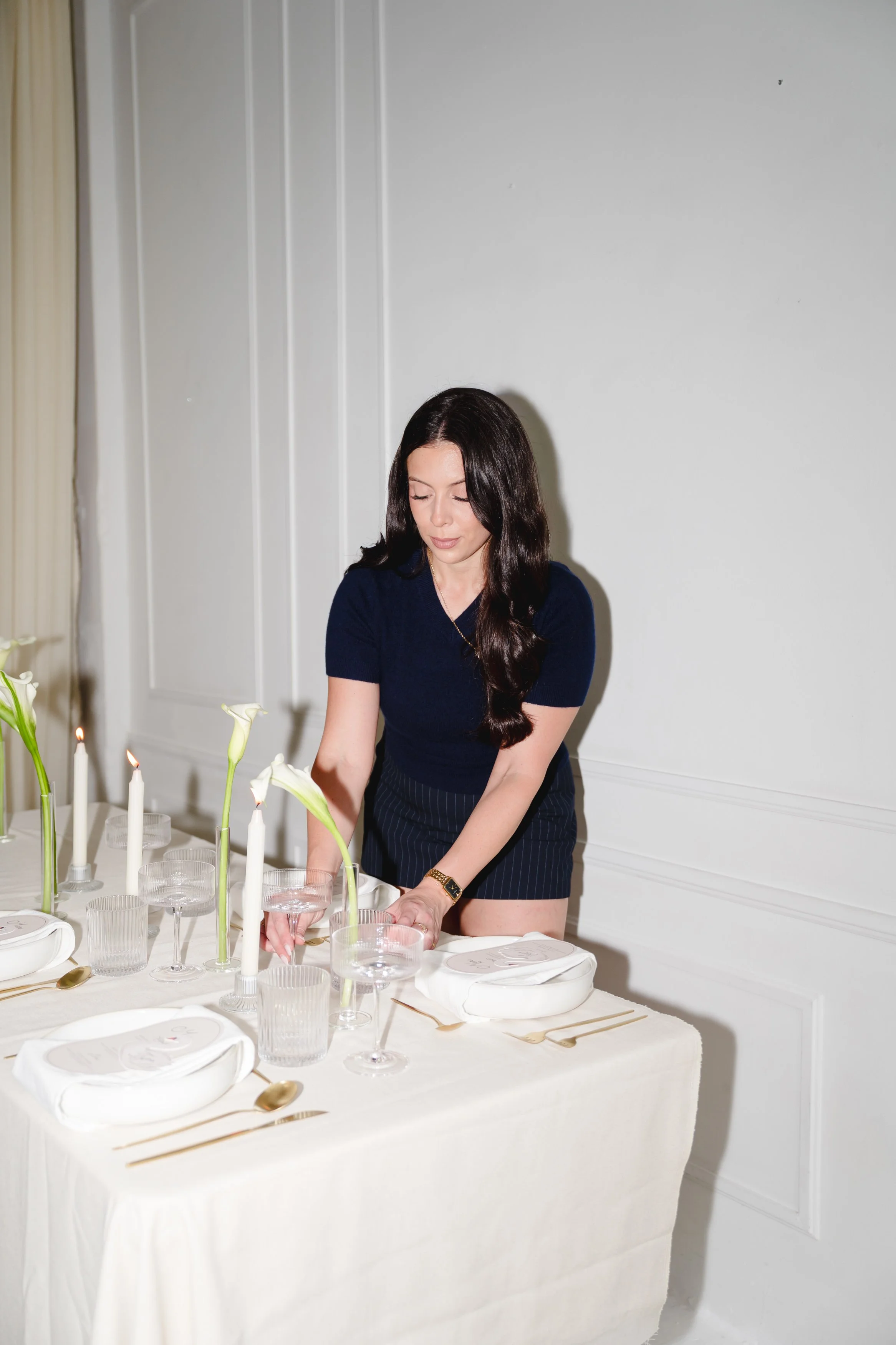 A woman with long dark hair in a navy blue shirt and striped shorts is setting a dining table with white plates, gold cutlery, clear glassware, and vases with white calla lilies. The table background is white with candles and a white wall.