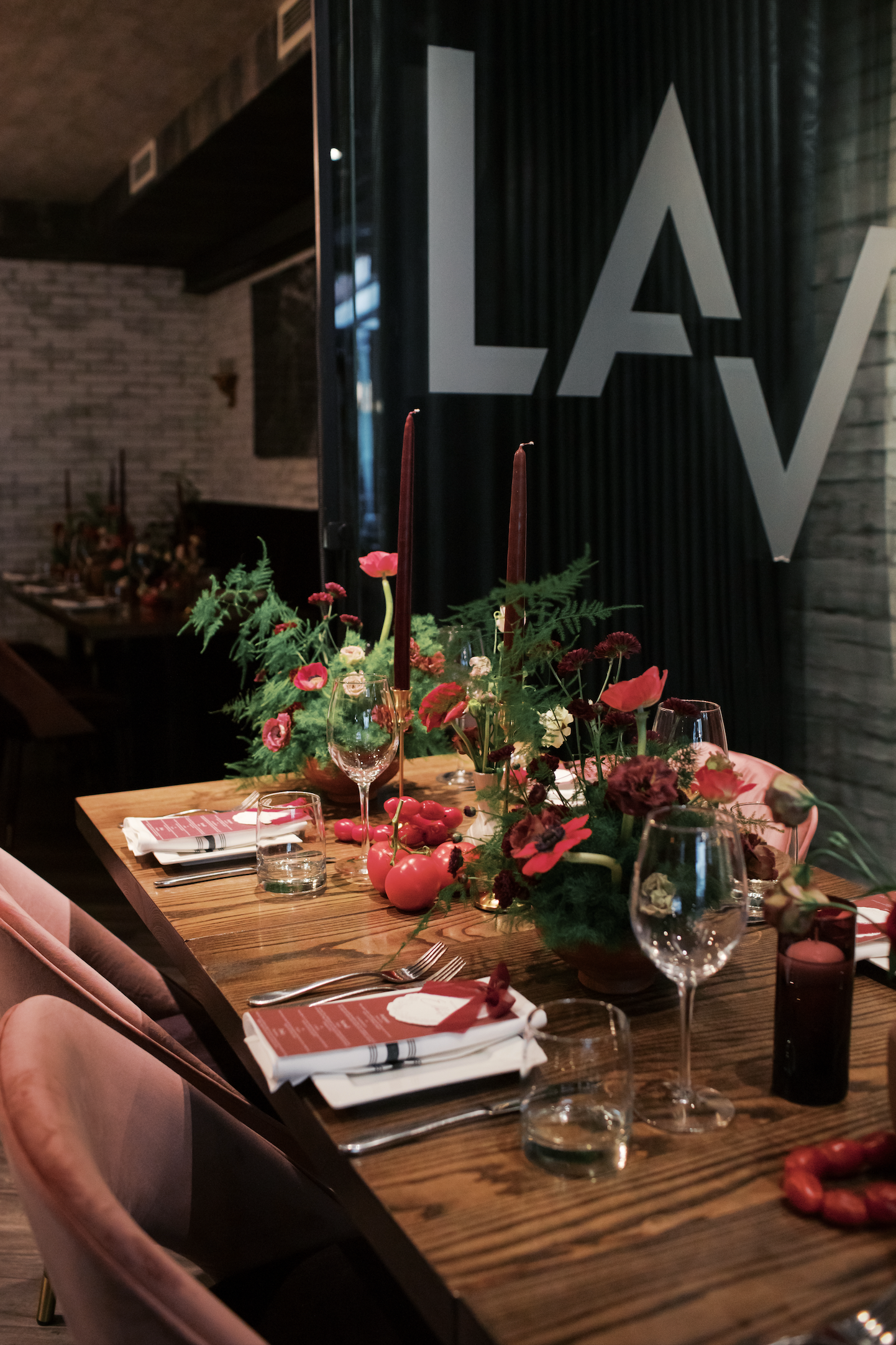 A decorated dining table with floral arrangements, red candles, wine glasses, and napkins, set for a formal meal in a restaurant with brick walls and a large black and white sign reading 'LA'.