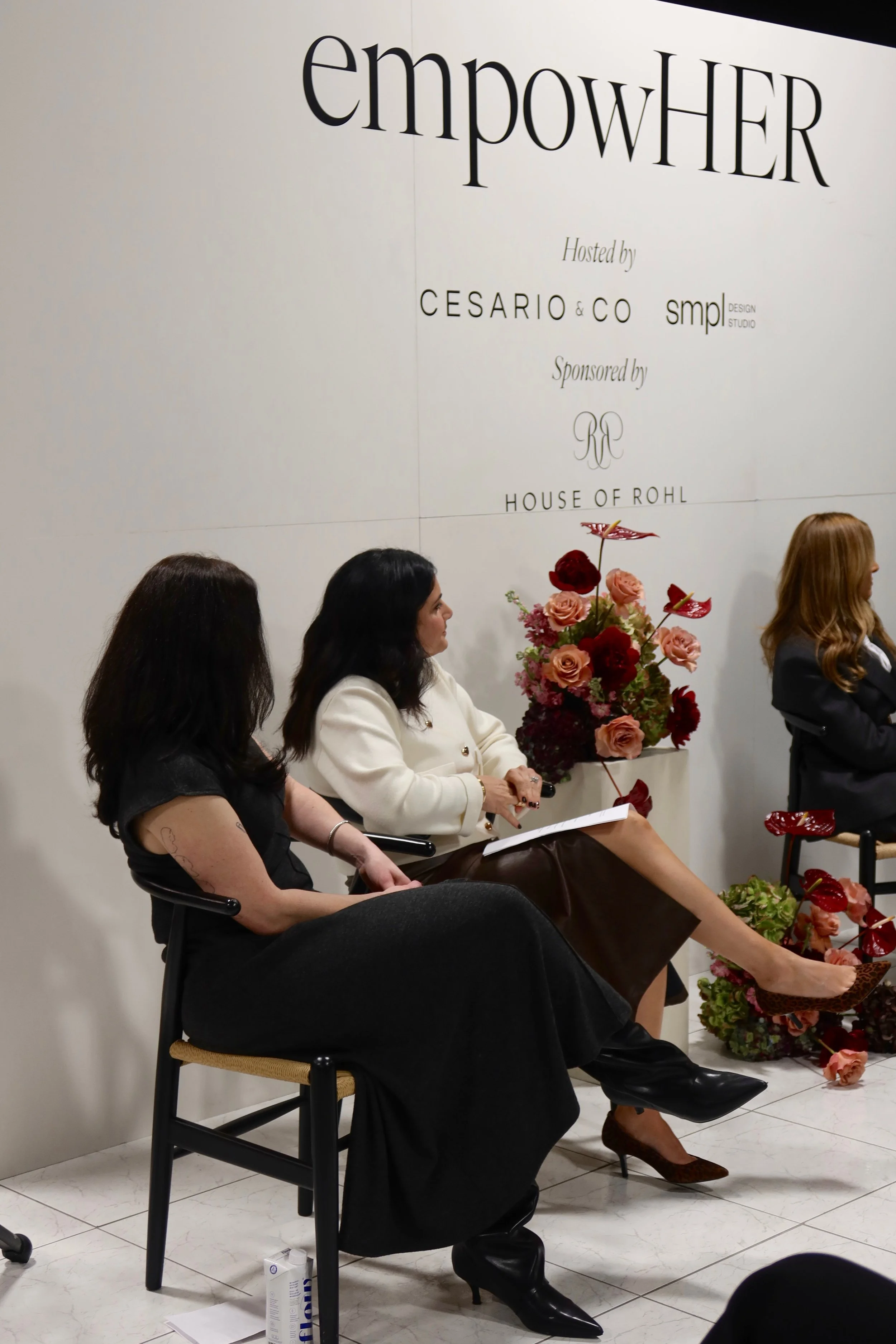 Three women sitting on chairs in front of a white wall with large text that reads 'empowHER' and smaller text mentioning hosts and sponsors. Two women are engaged in conversation, with one holding a paper. There are flower arrangements on the floor and a small table nearby.