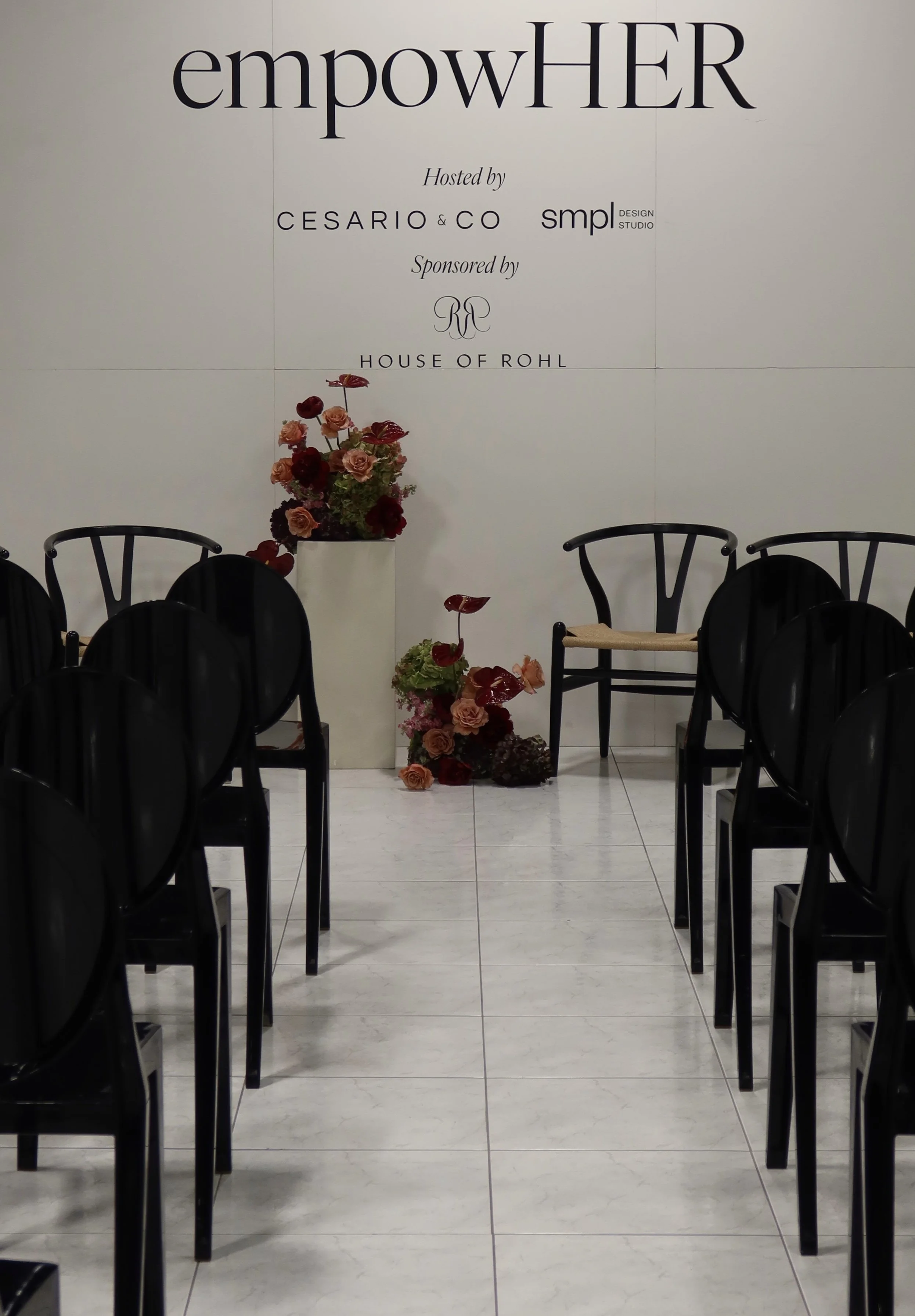 Event space with white walls and a white tile floor, decorated with a tall floral arrangement of pink and red flowers. Black chairs are arranged around the room, and a large sign on the wall reads 'empowHER' with the words 'Hosted by CESARIO & CO' an