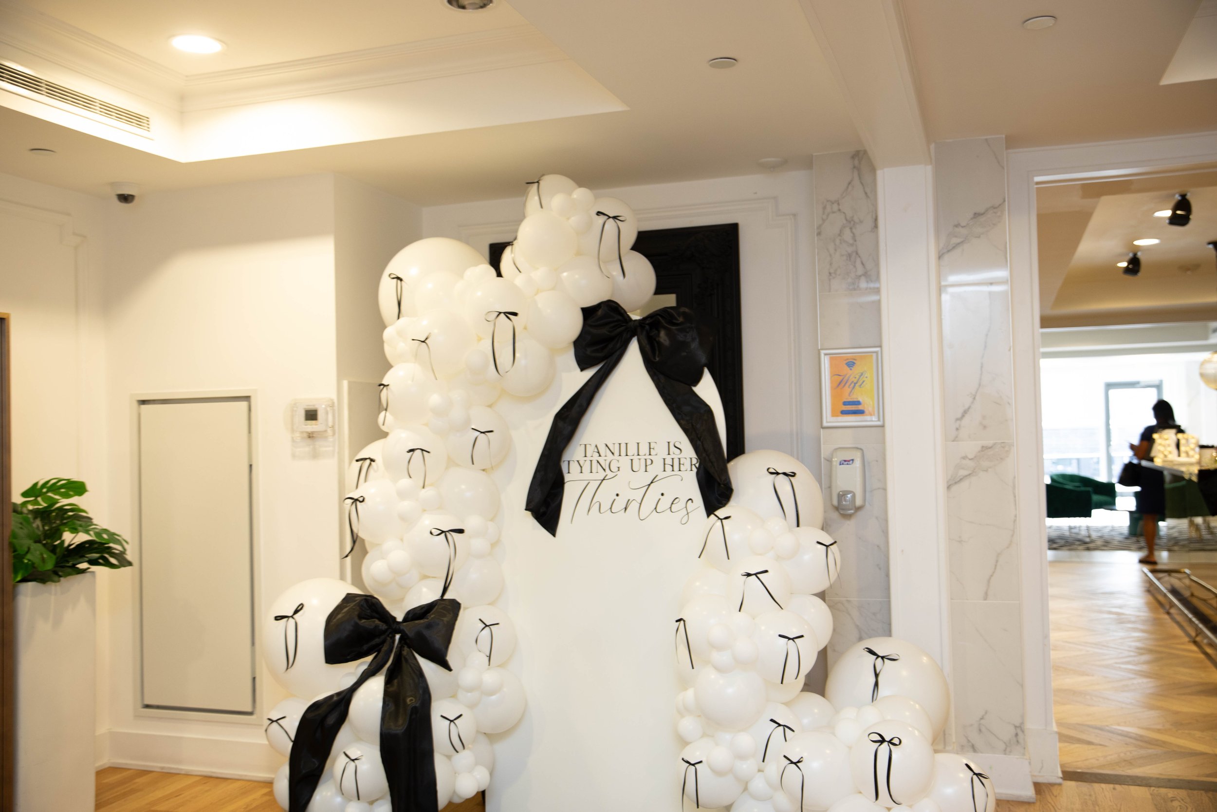 A balloon display featuring white balloons tied with black ribbons and large black bows, with a sign that reads 'TANILLE IS Tying Up Her Thirties' in a decorated indoor space.
