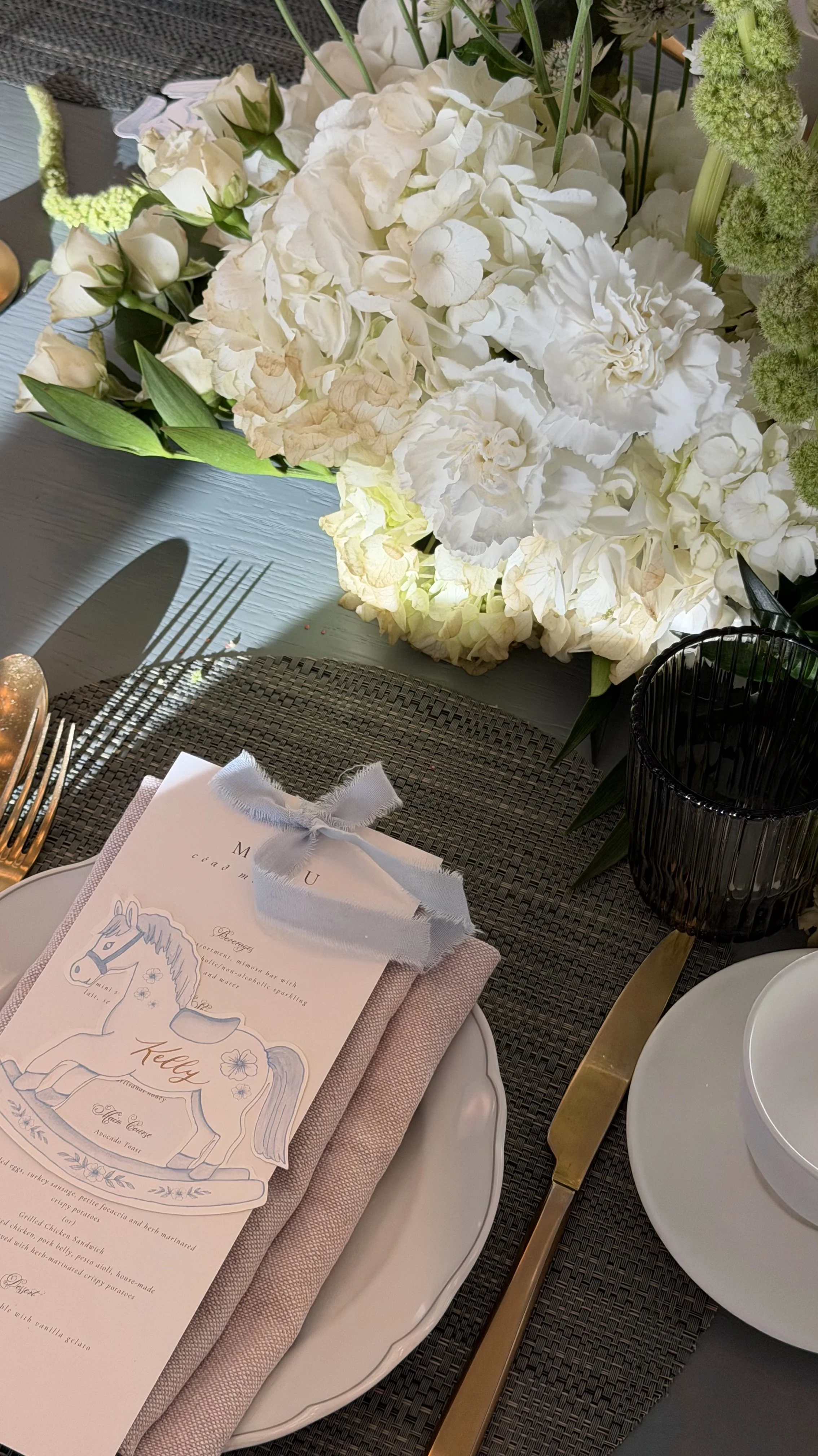 A beautifully set table with a large bouquet of white flowers, a menu with a unicorn illustration, pink and white napkins, and gold-cutlery, including a knife, on a dark woven placemat.