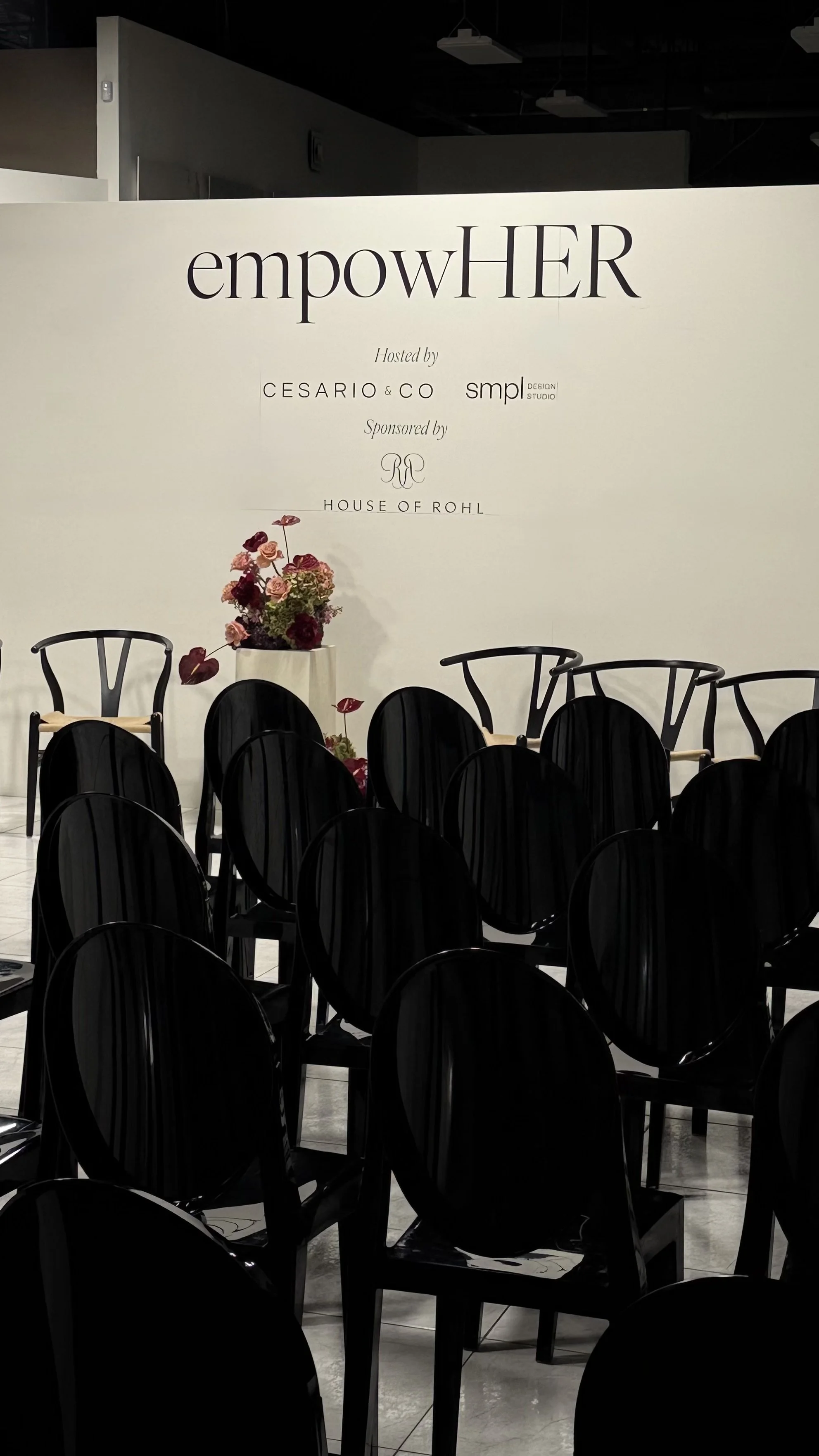 Event space with black chairs arranged in rows, a white wall with the words 'empowHER' and event details, and a floral arrangement in a white vase.