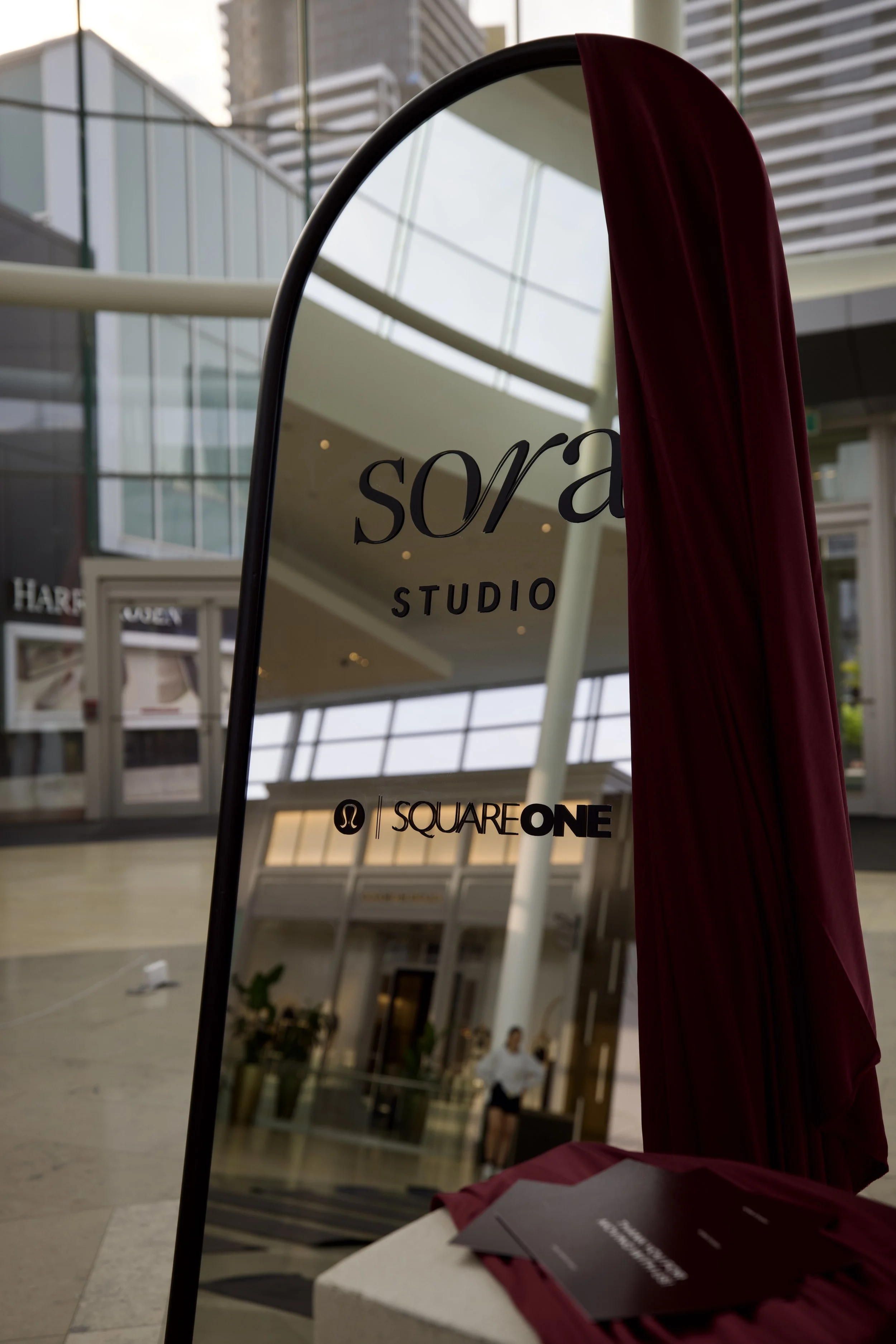 Mirror with the logo 'sora STUDIOS' and 'SQUAREONE' at a shopping mall, with a red curtain partially covering the mirror, and brochures placed on a table in the foreground.