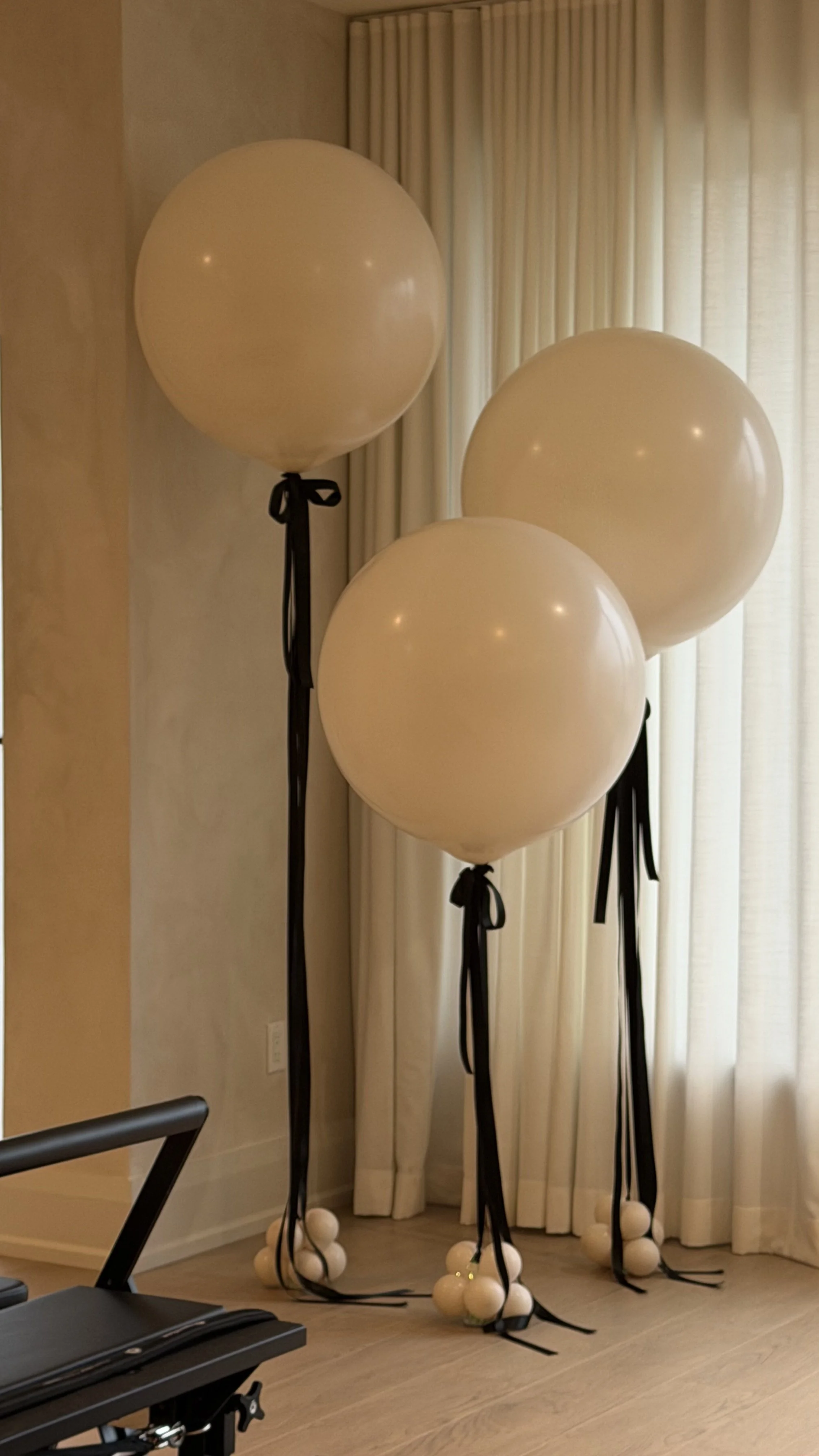 White balloons with black ribbons tied around the bases, standing on black stands with ball-shaped bases, set against cream-colored curtains and wall.