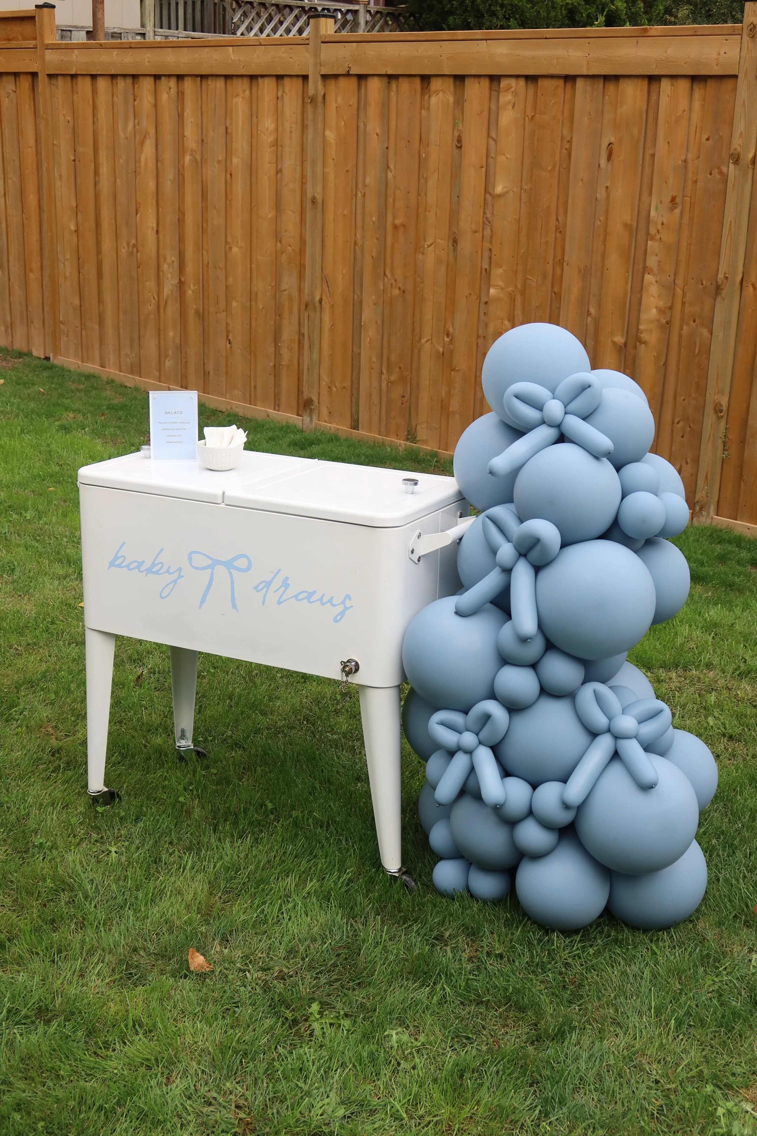 A white refrigerated drink table with the text 'baby R draw' on it, decorated with a large blue balloon sculpture resembling a flower or abstract shape, positioned outdoors on a grassy lawn with a wooden fence in the background.