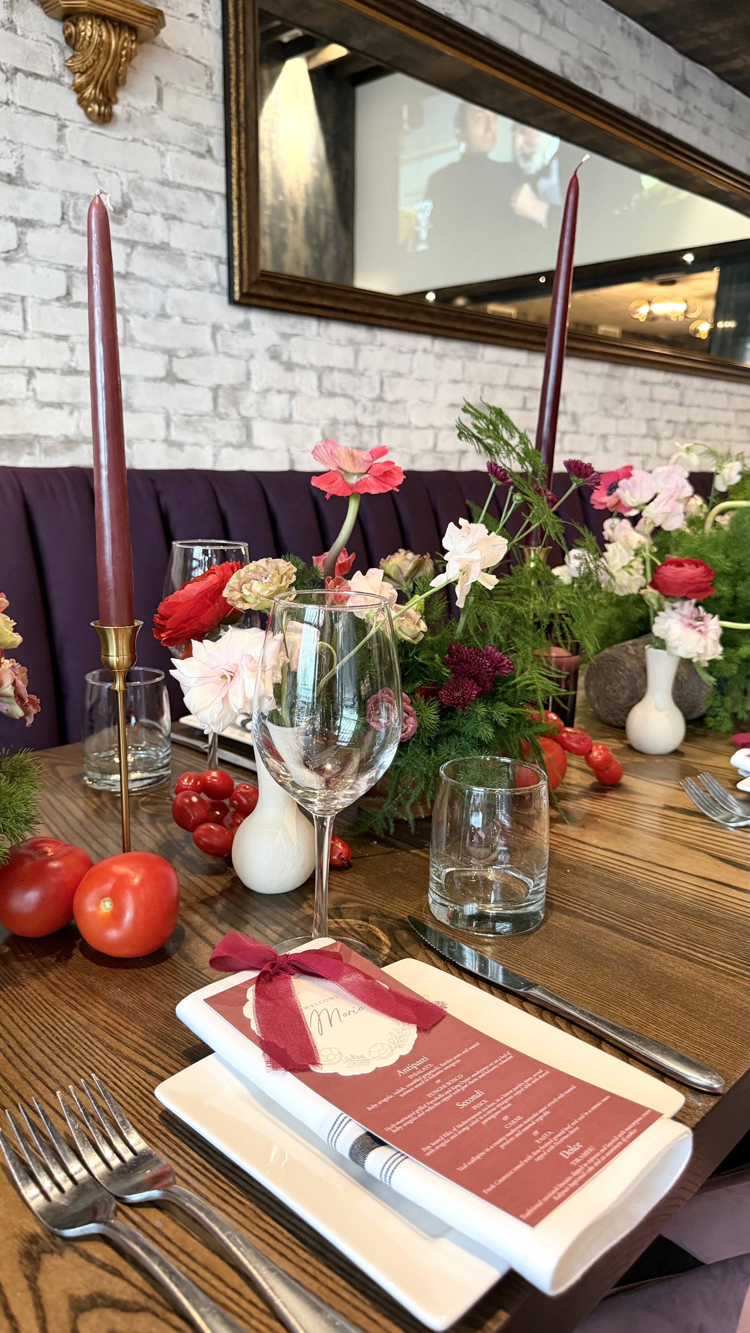 A dining table decorated with flowers, tomatoes, and candles, with a menu on a white plate, set with silverware.