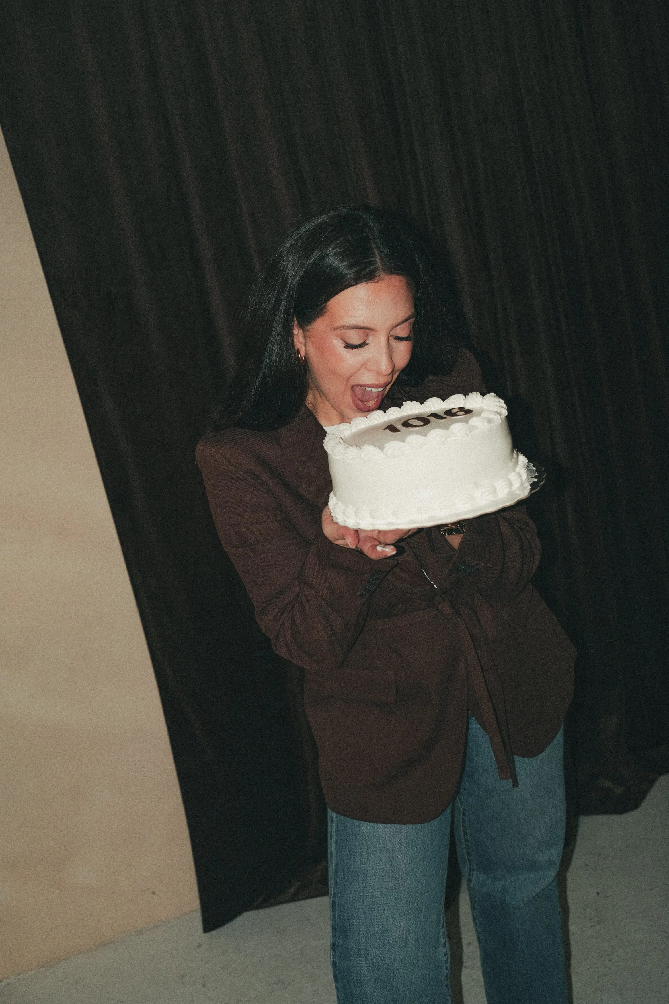 A woman with black hair wearing a brown blazer and blue jeans is holding a white cake with the number 10 on top, celebrating an anniversary or milestone.