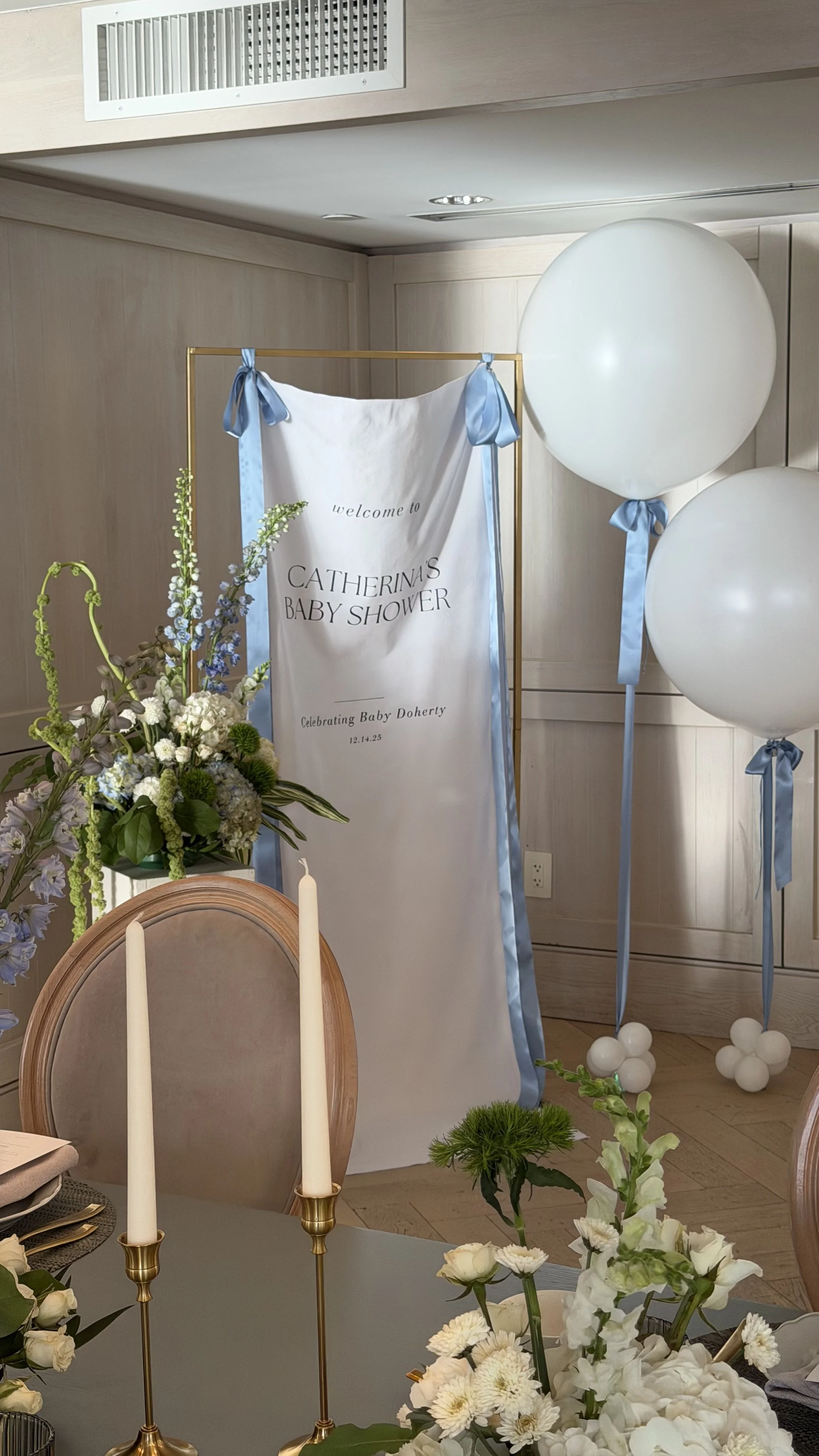 Decorations for a baby shower, including white balloons with blue ribbons, a floral arrangement with white and purple flowers, a welcome sign with blue ribbons, and a table with candles and plates.