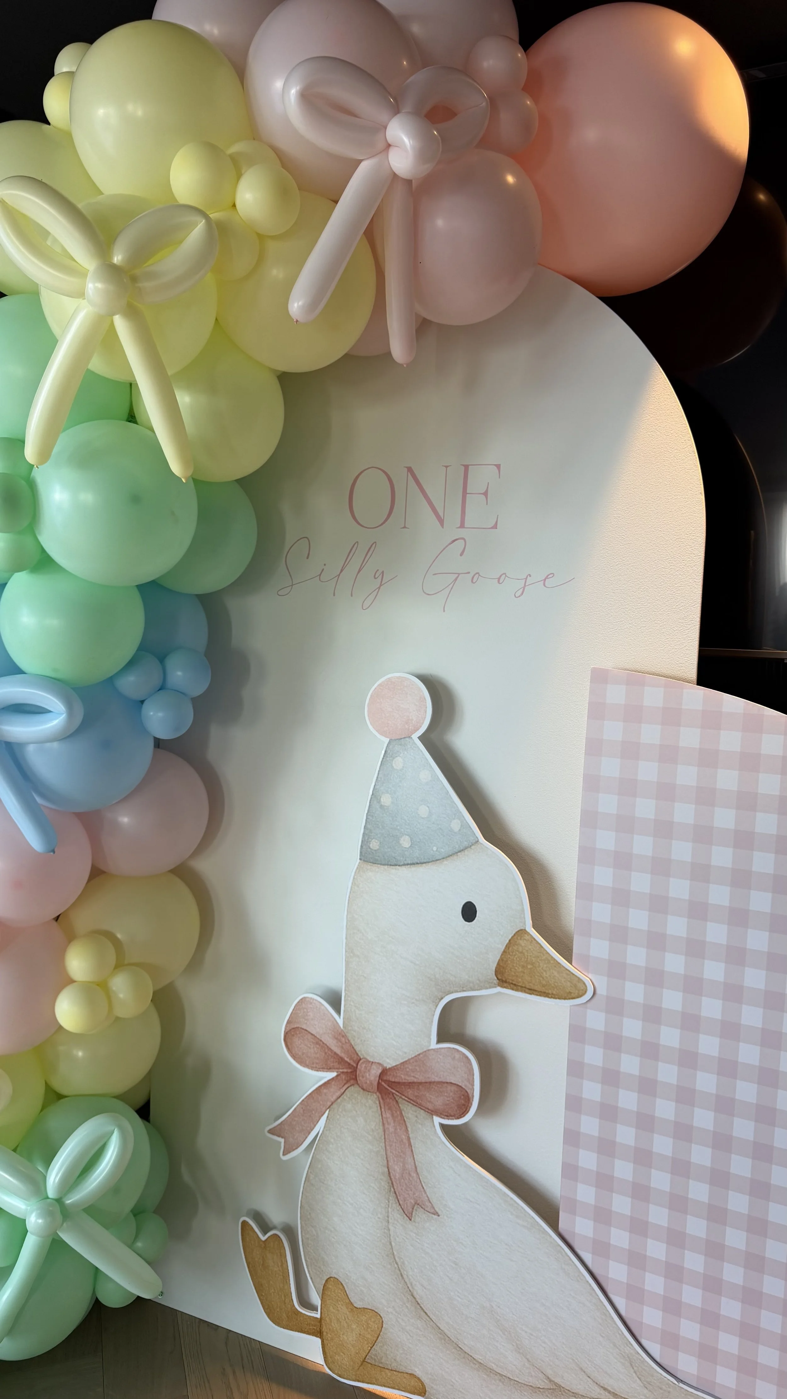 A decoration with pastel-colored balloons and a cartoon-like paper cutout of a goose wearing a party hat and bow, with the words "ONE Silly Goose" written on a sign.