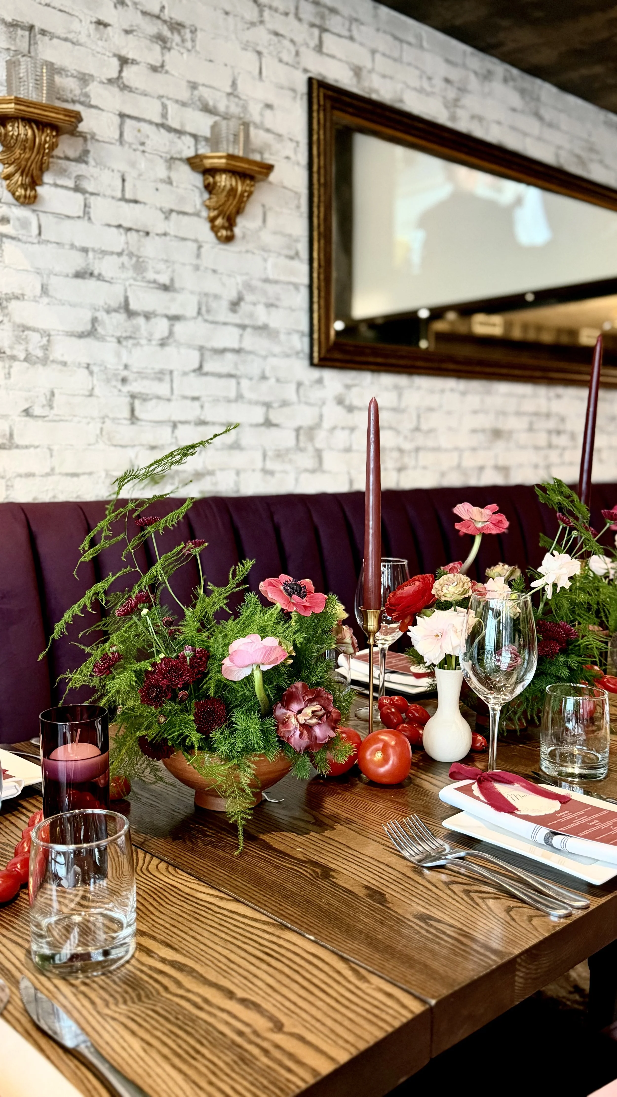 A decorated dining table with a floral centerpiece, pink and red flowers, candles, glasses, and cutlery in a restaurant setting with a white brick wall and framed mirror in the background.
