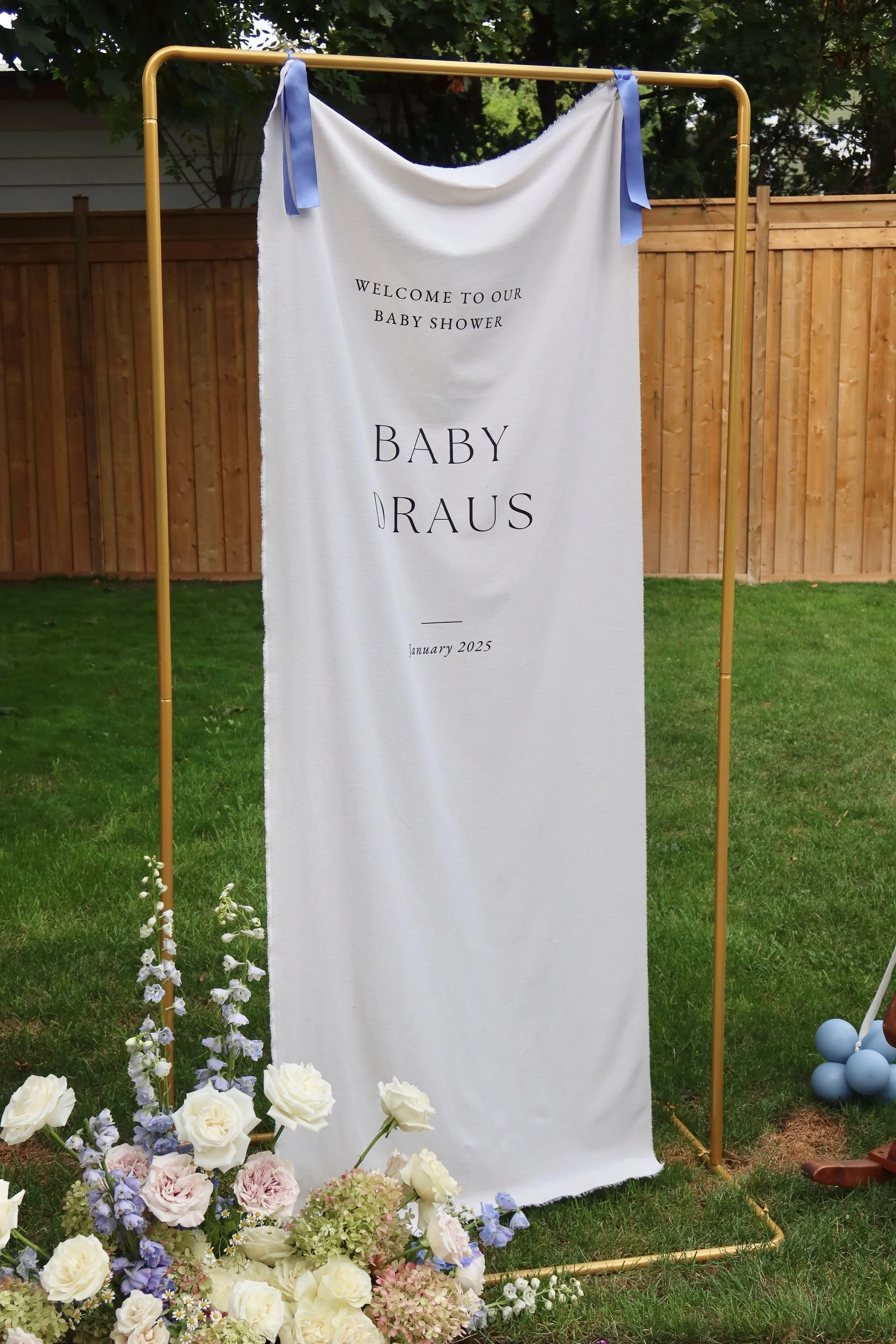A white fabric banner with black text that reads "Welcome to our baby shower, Baby DRAUS, January 2025" hangs on a gold frame outdoors, with a wooden fence and trees in the background, and floral arrangements including white roses, hydrangeas, and pu