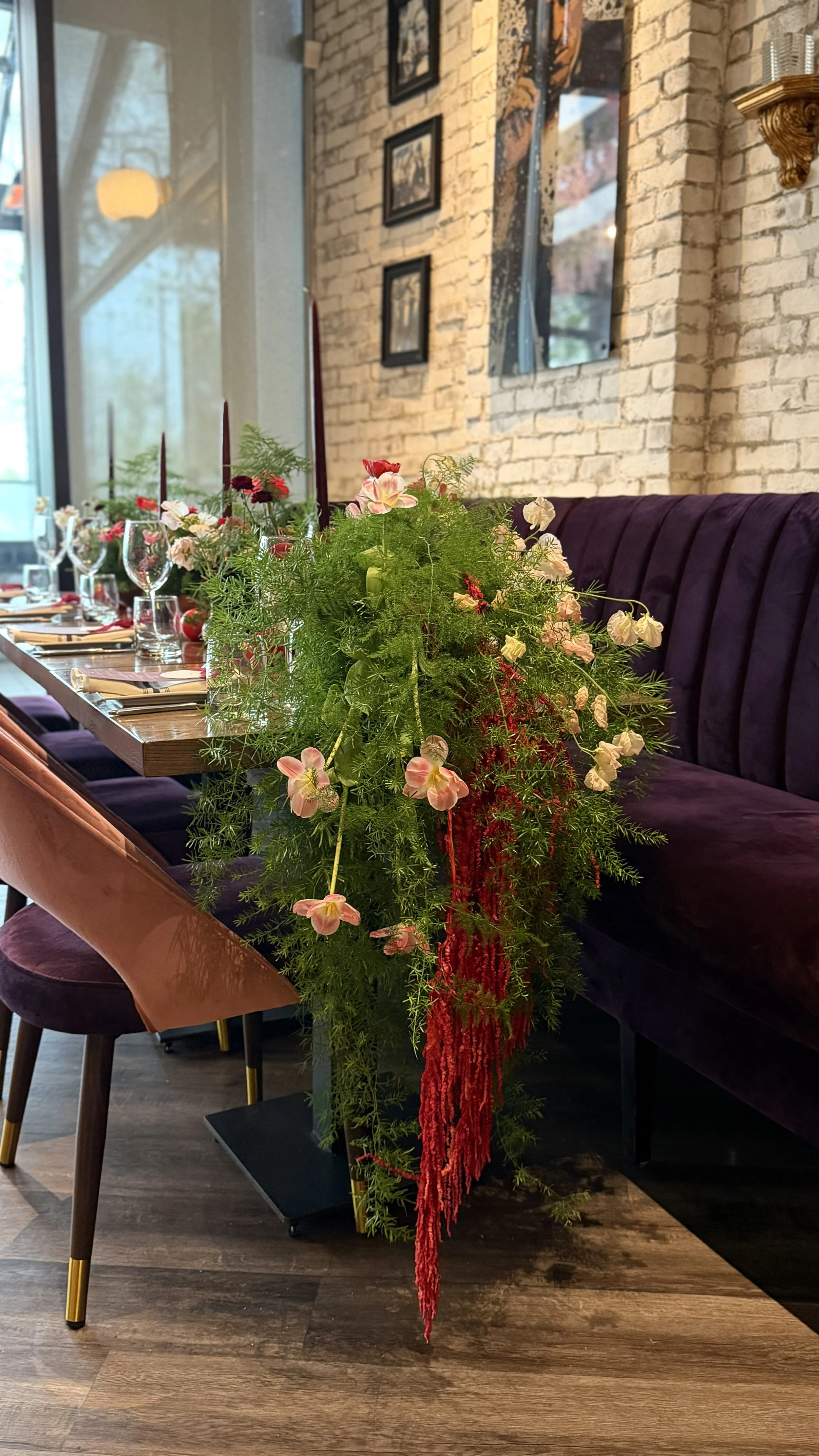A long dining table with purple velvet chairs and a floral centerpiece with pink, red, and white flowers and green foliage in a restaurant with brick walls and framed pictures.