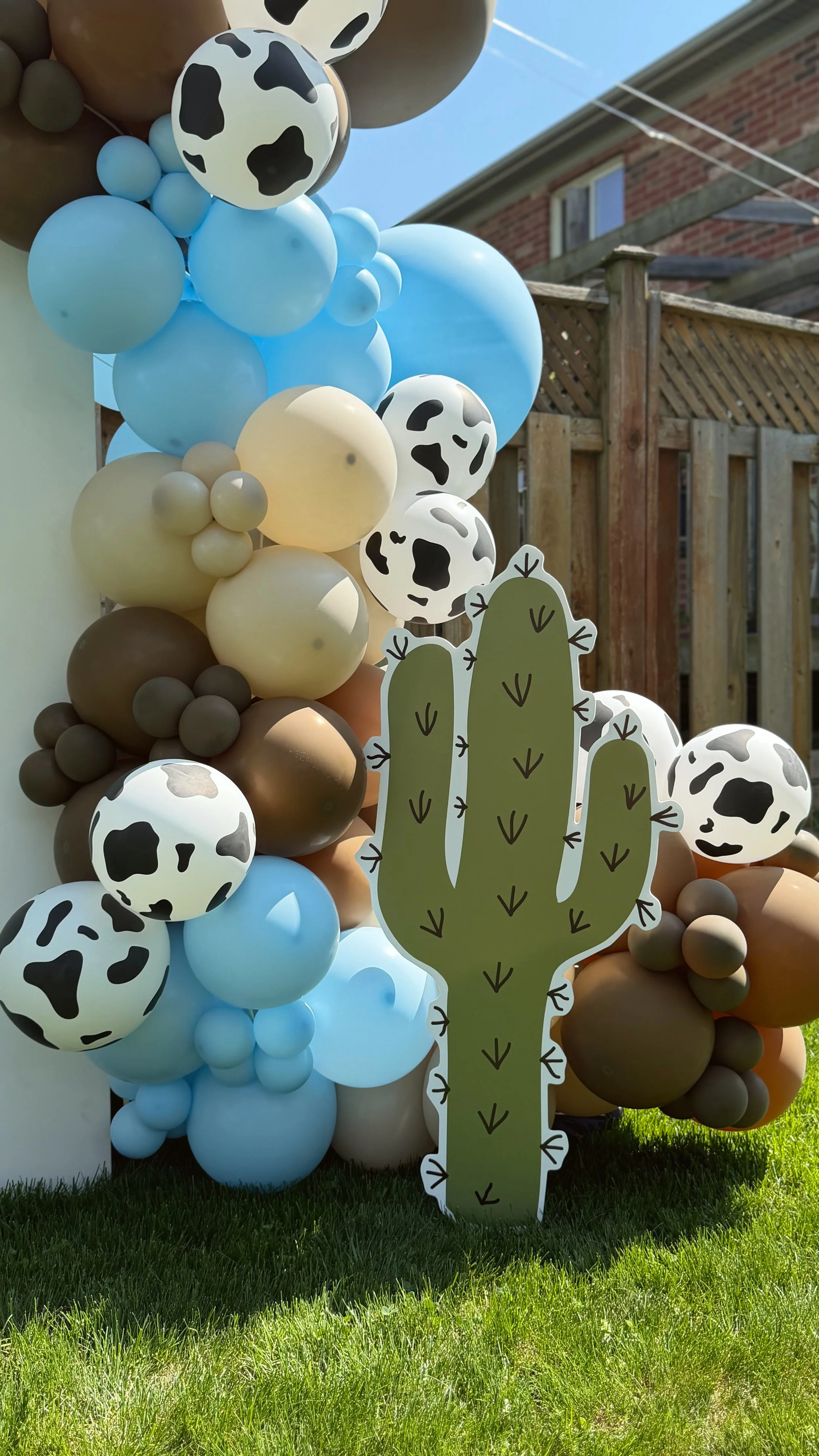A decorative balloon arrangement with tiger print, solid, and pastel balloons, featuring a large paper cactus cutout attached to the display outdoors on a lawn.