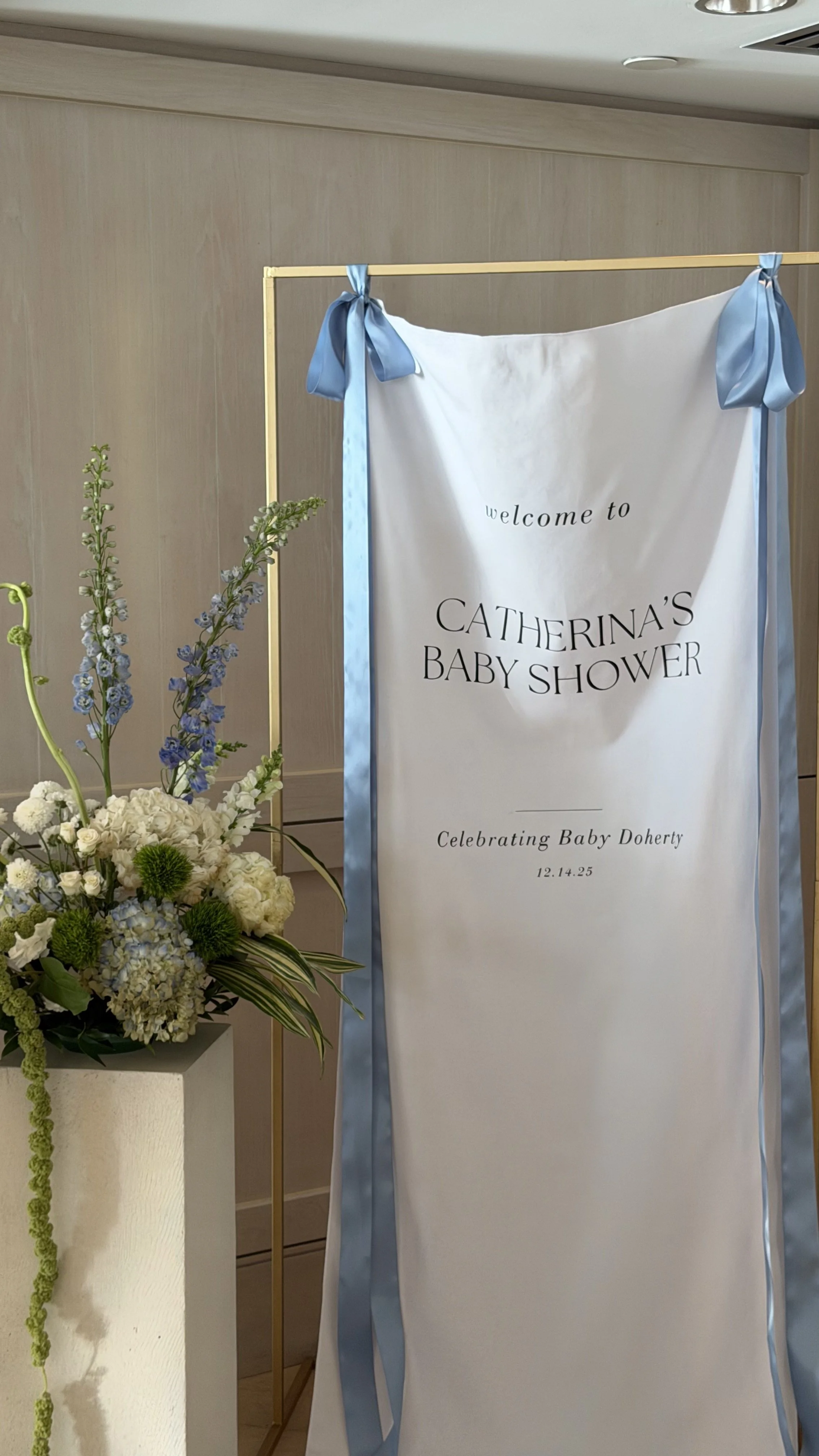 A welcome sign for Catherina's Baby Shower with blue ribbons and a floral arrangement nearby, celebrating Baby Doherty on December 14, 2025.