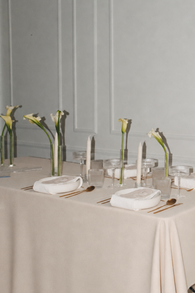 Elegant dining table with white calla lilies, candles, glassware, cloth napkins, and gold cutlery in a sophisticated setting.