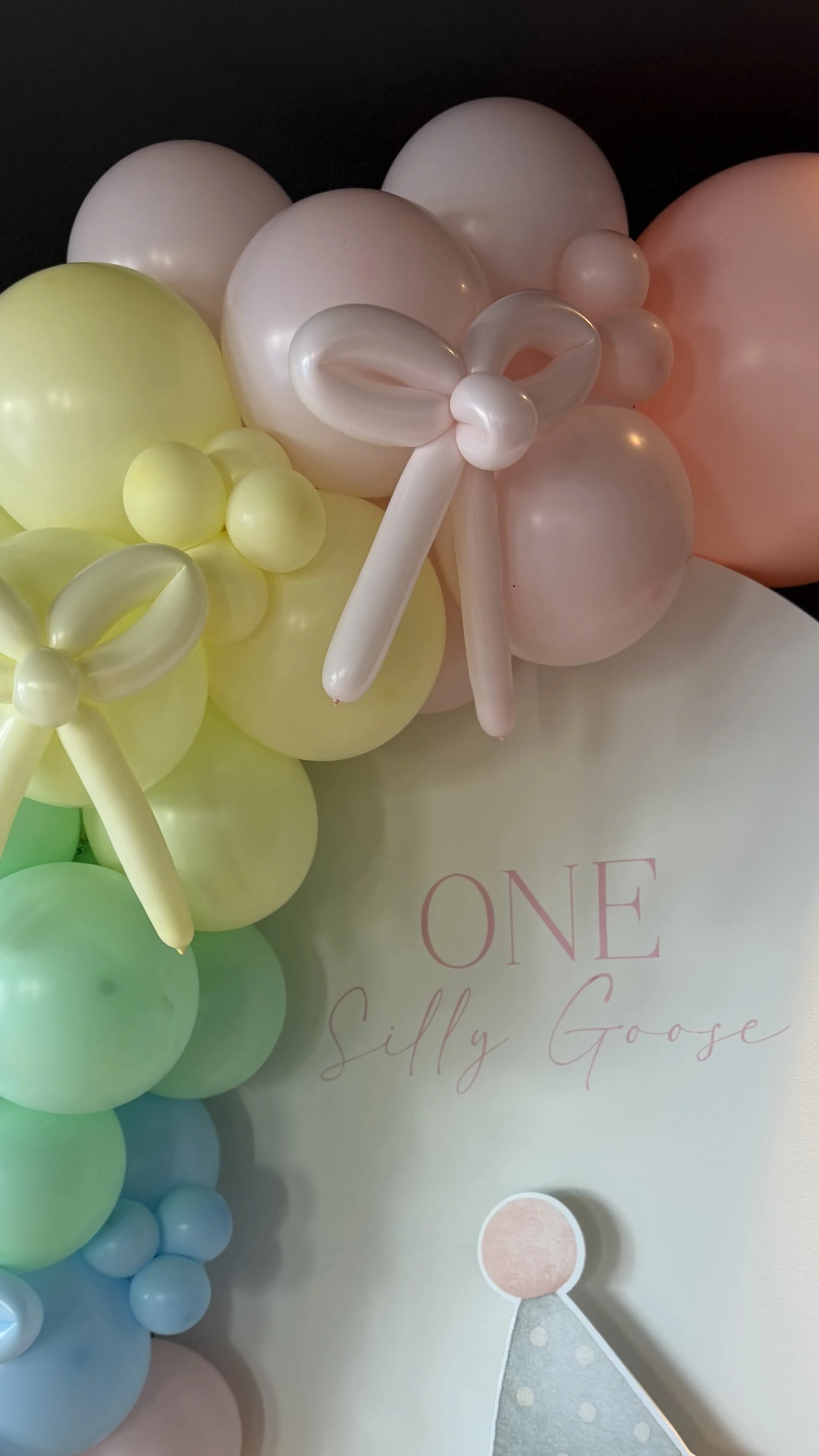 Colorful balloon decorations with pastel green, yellow, pink, blue, and peach balloons arranged around a sign that says "ONE Silly Goose" and a cutout of a party hat figure.