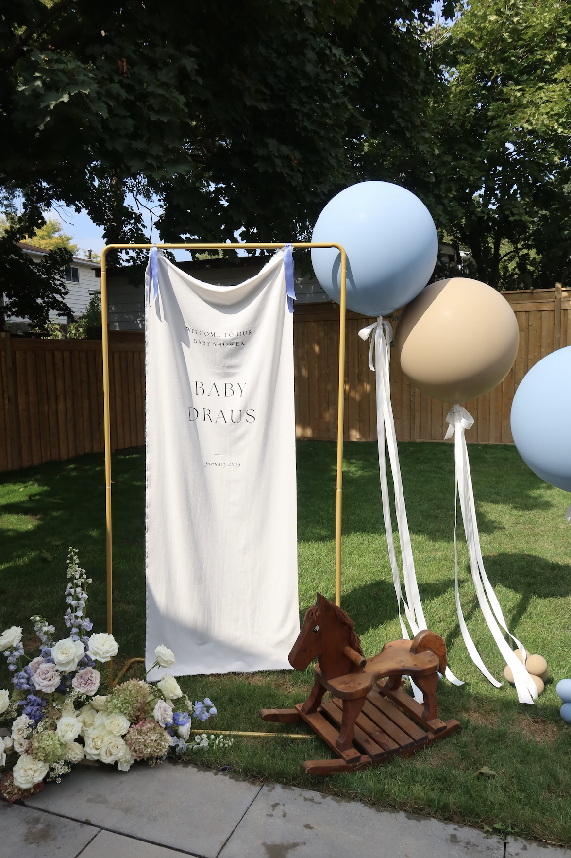 Outdoor baby shower decoration with large blue and beige balloons tied with white ribbons, a white fabric banner reading 'Welcome to our baby shower, Baby Draus, January 2025' hanging from a yellow frame, a floral arrangement of white and pastel flowers on the ground, a small wooden rocking horse, and a backyard with green grass, trees, and a wooden fence in the background.