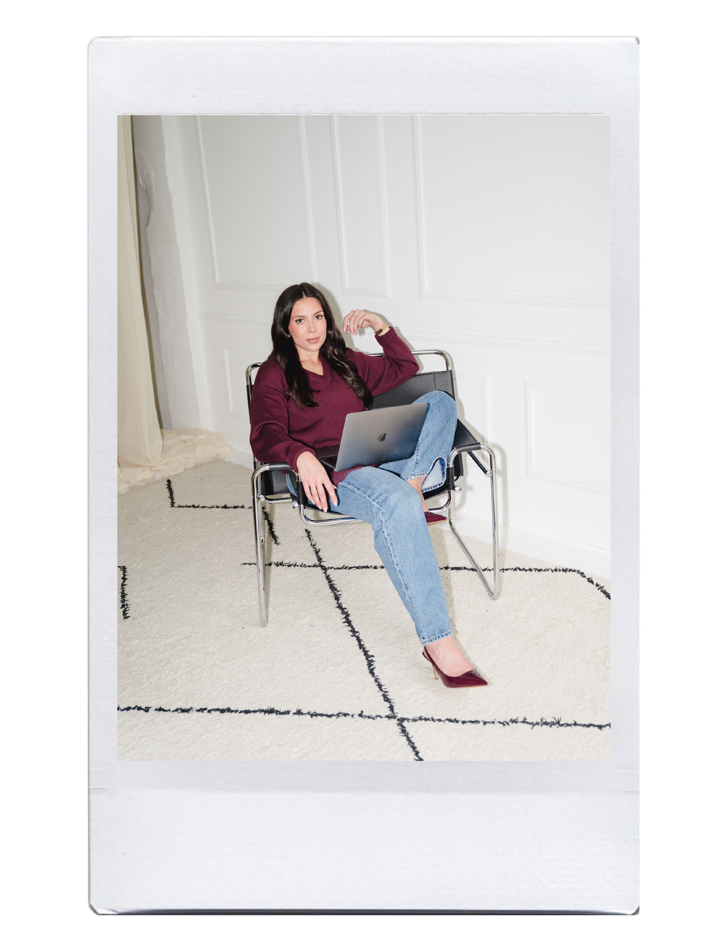 A woman with dark hair sitting in a black chair, wearing a maroon sweater, blue jeans, and high heels, with a laptop on her lap, in a room with white walls and a white carpet.