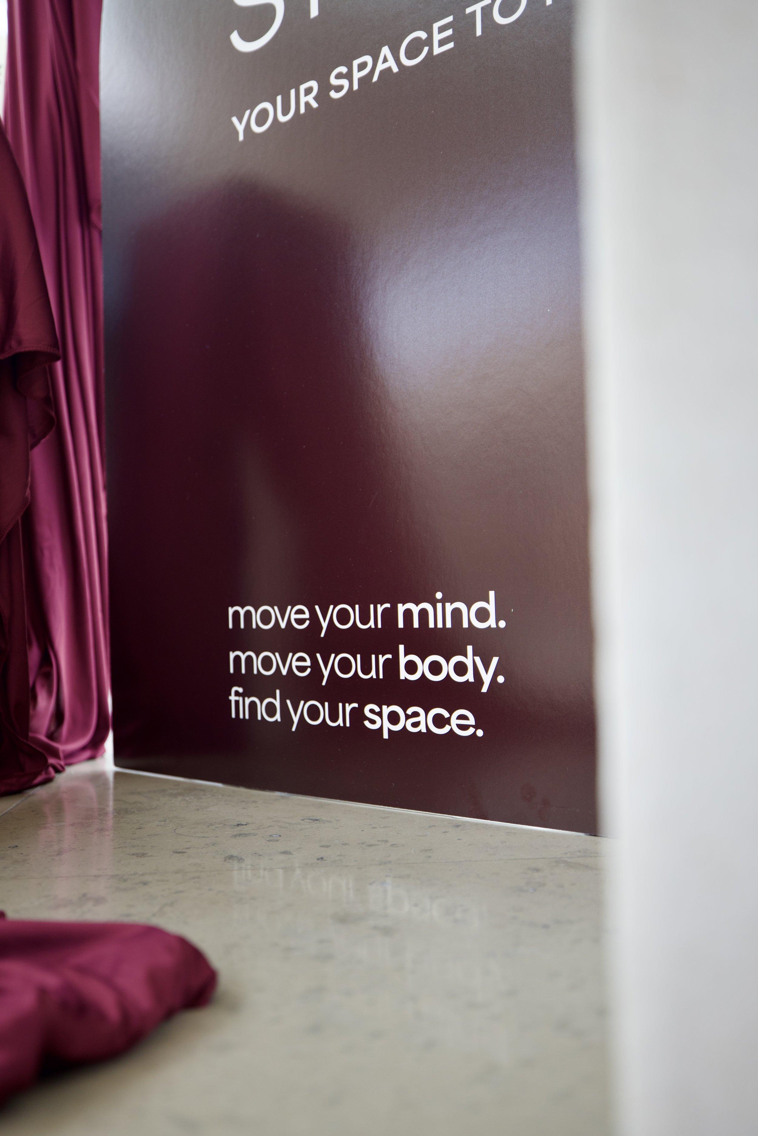 Close-up of a wall with motivational text that says: "move your mind, move your body, find your space". Part of a pink fabric or curtain is seen on the left side of the image and a pink object, possibly a bag, is on the ground in the foreground.