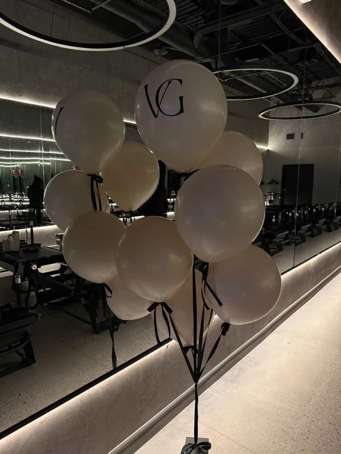 A bunch of white balloons with black ribbons tied to them, displayed in front of a large mirror in what appears to be a fitness or pilates studio with exercise equipment and mirrors on the walls.