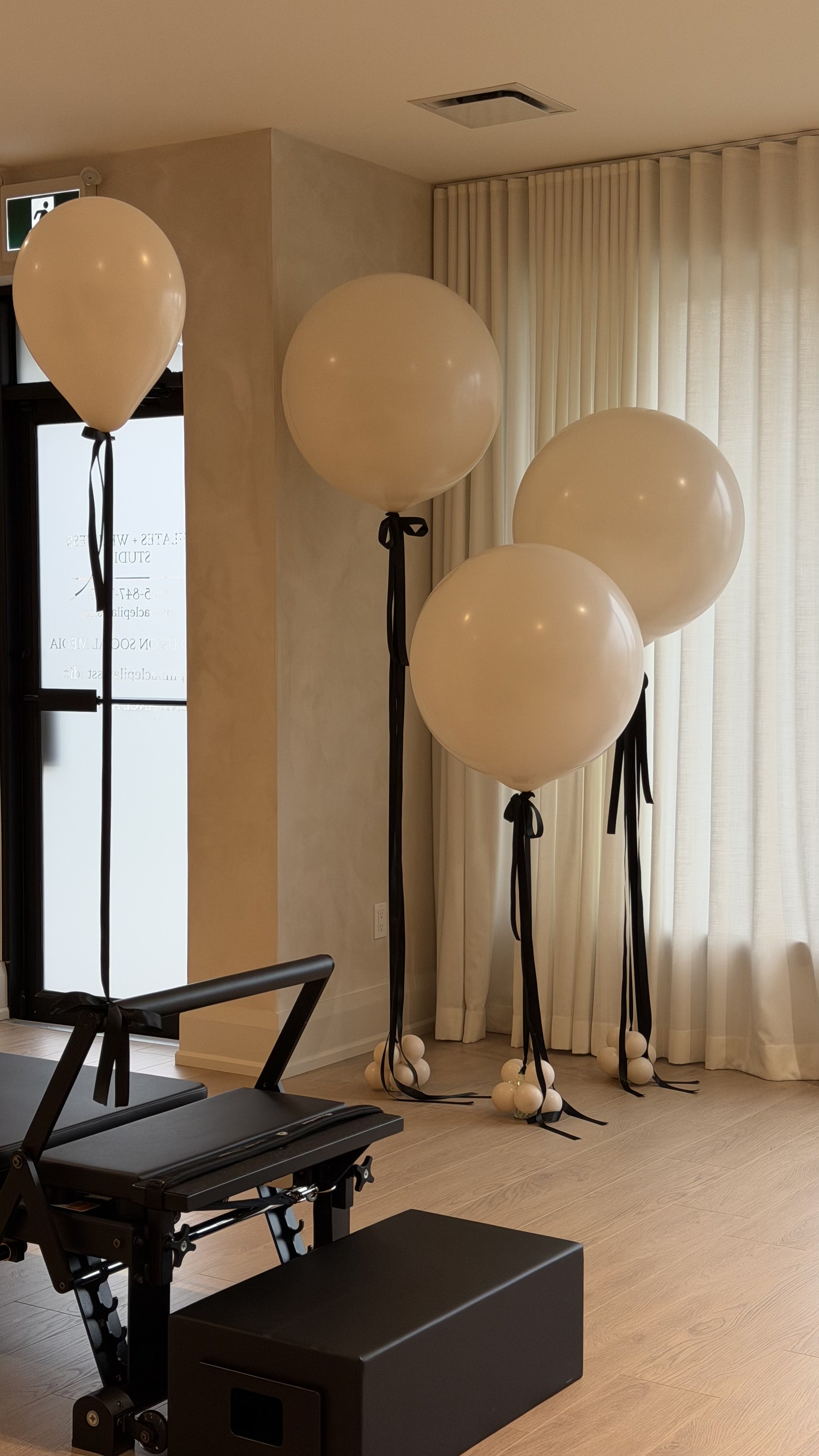 Four white balloons tied with black ribbons and decorated with white balls at the bottom, standing indoors near beige curtains.
