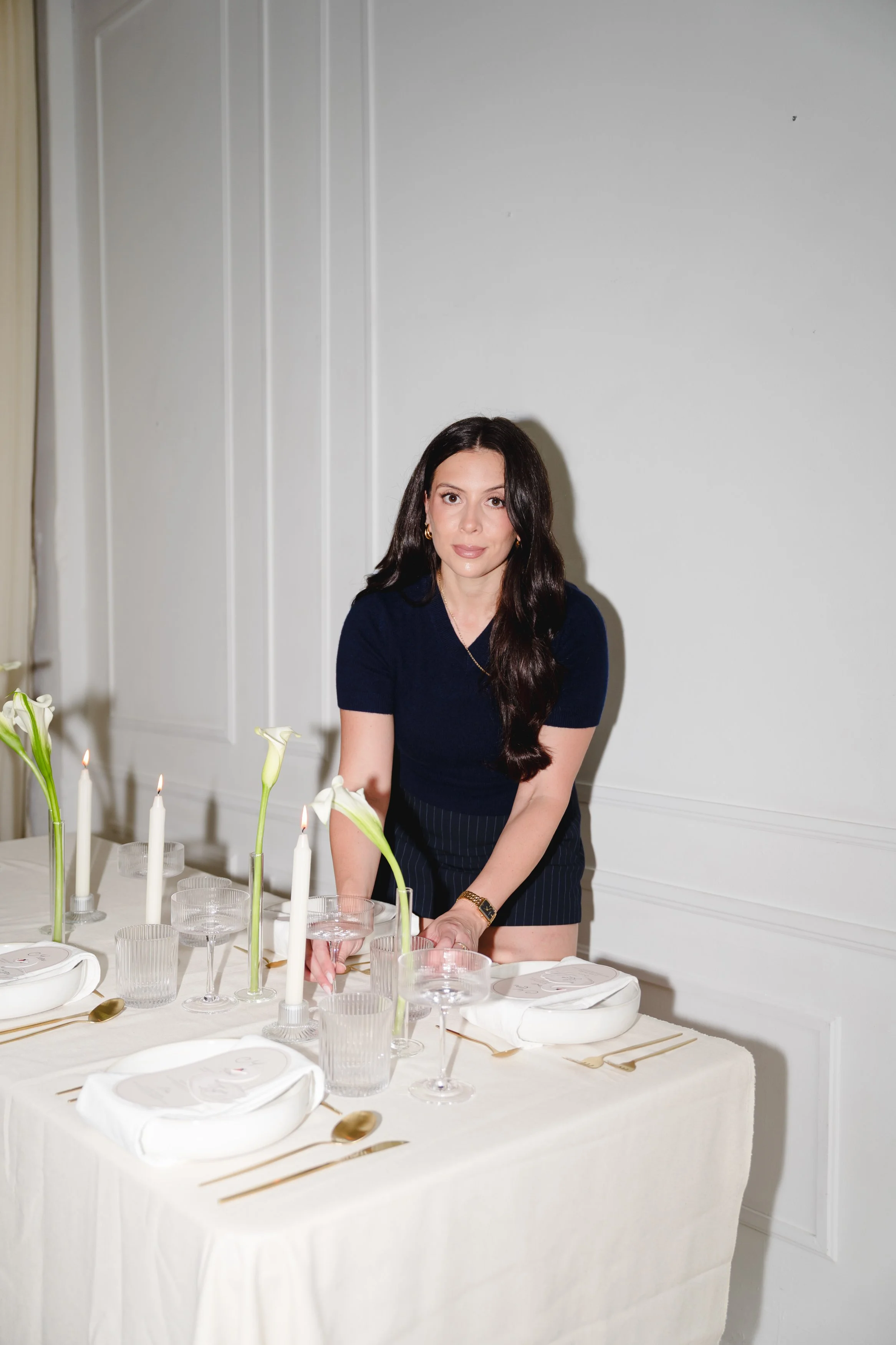 A woman with long dark hair sitting at a decorated dining table with white plates, gold cutlery, and white floral centerpieces with lit candles, in a room with white walls.