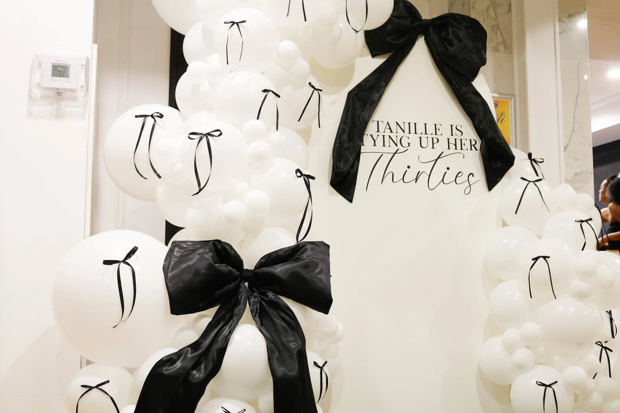 Decorative display of white balloons with black ribbons and bows, along with a large black bow. A sign reads 'TANILLE IS TYING UP HER Thirties' in elegant black script. The setup is likely for a birthday celebration.