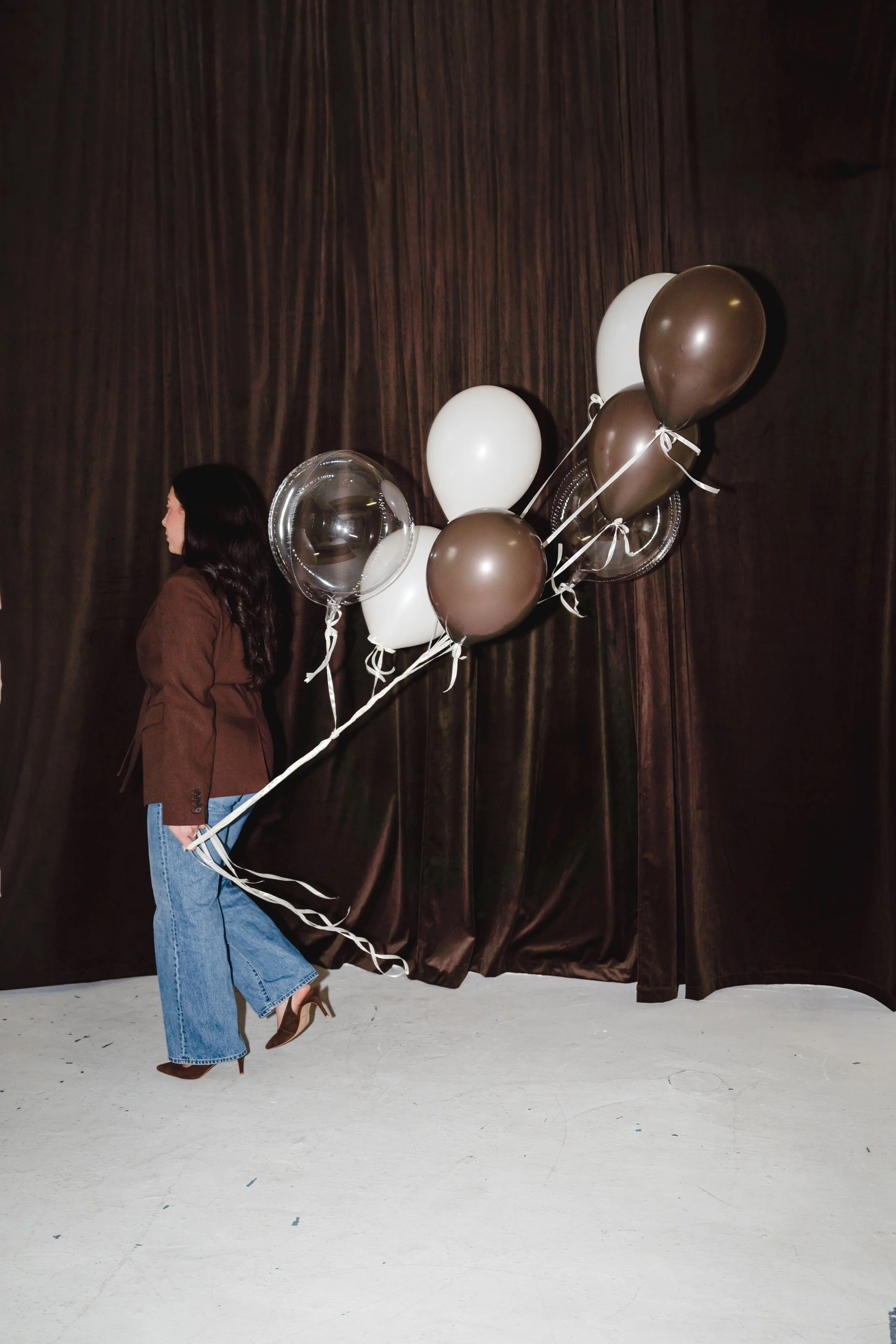 A woman with dark hair wearing a brown blazer, jeans, and high heels is holding a bunch of gold, silver, and white balloons. She is standing against a dark curtain background.