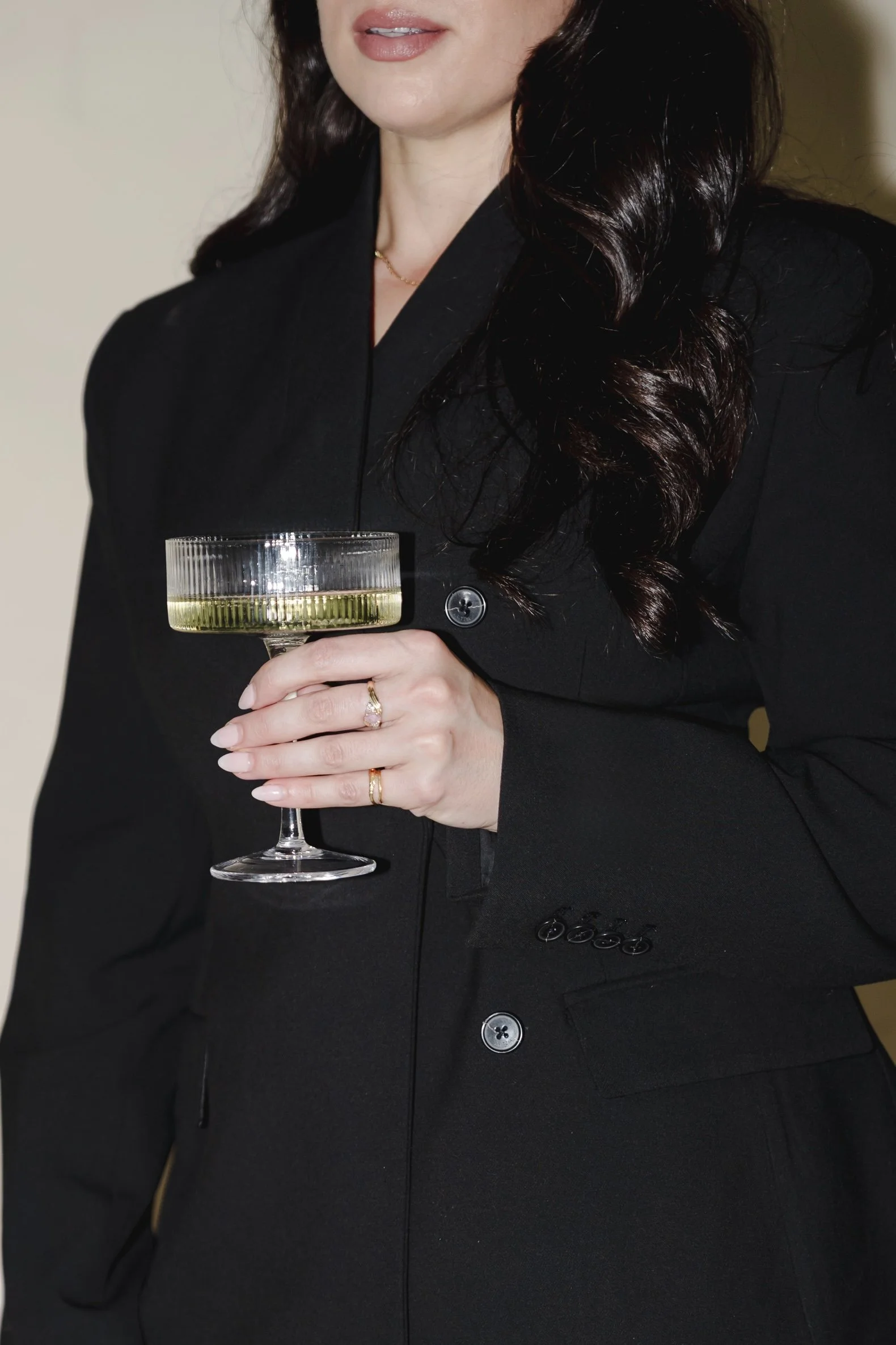 A woman dressed in a formal black blazer, holding a glass of white wine, with dark, wavy hair and wearing rings and a delicate necklace.