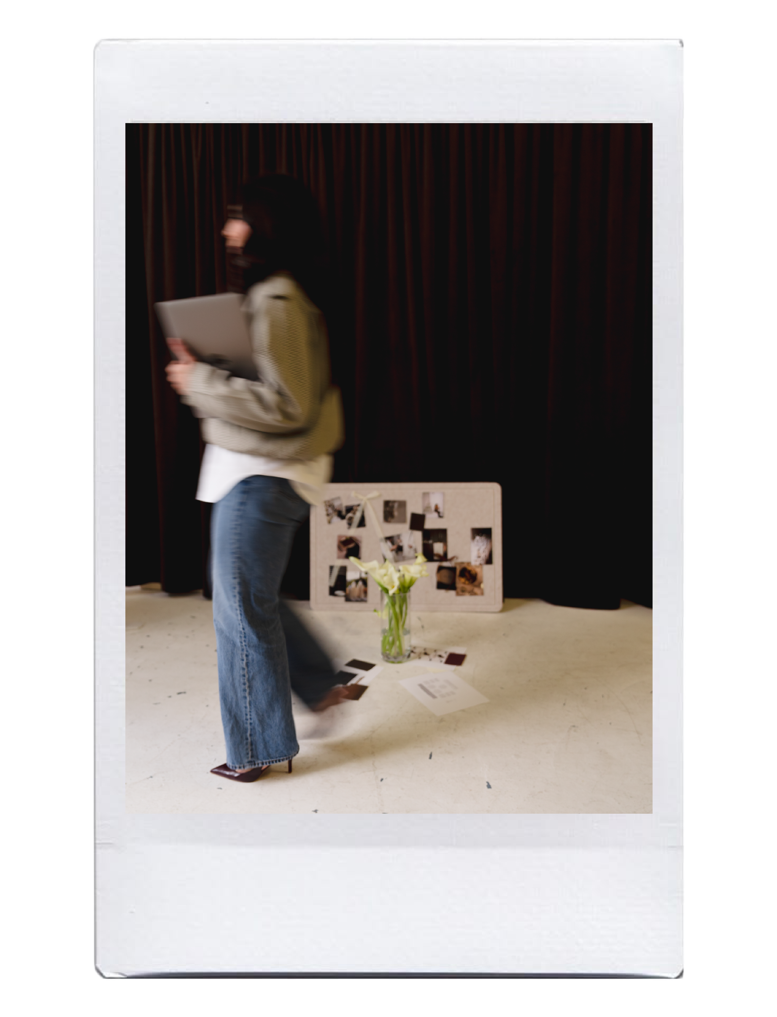 A woman in jeans and heels holding a laptop, walking past a whiteboard with photos and flowers in a vase in front of a dark curtain background.